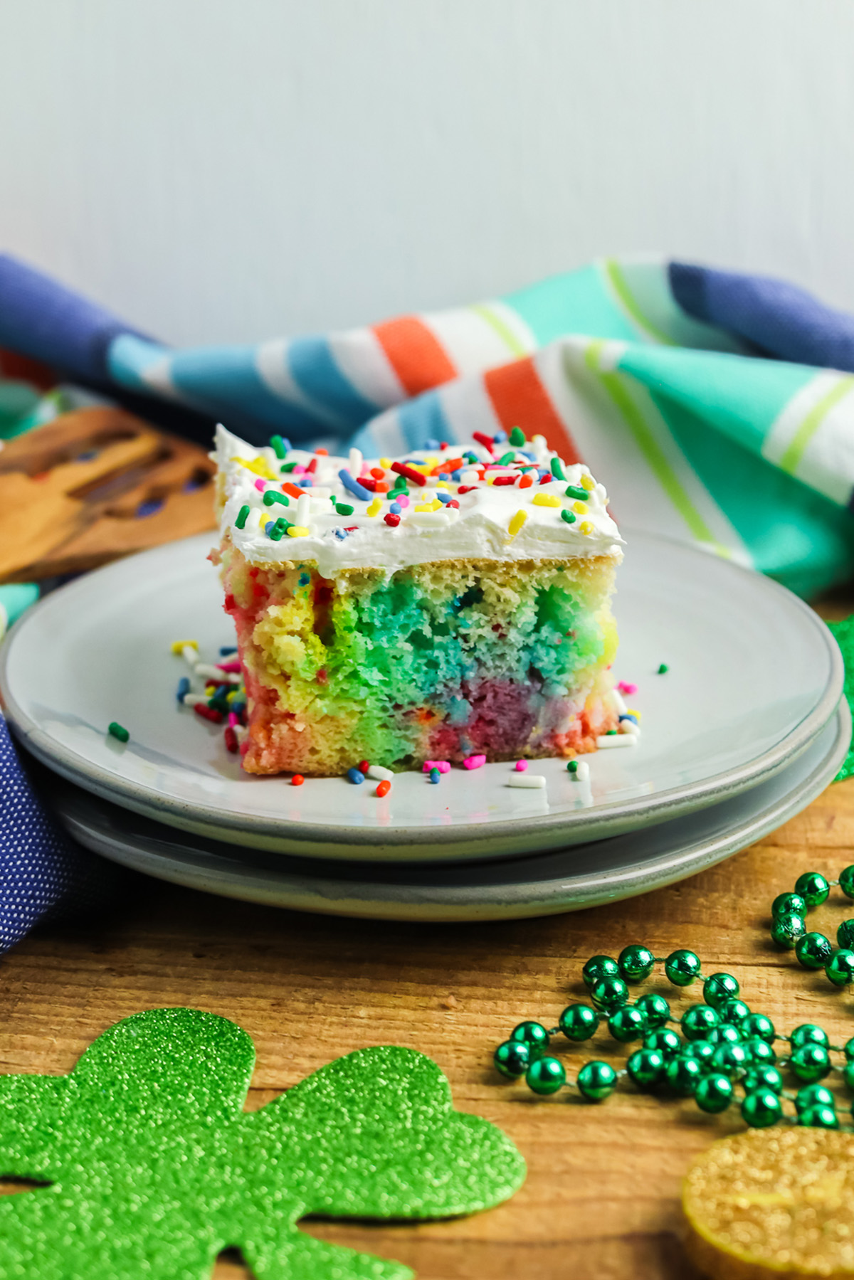 Slice of rainbow poke cake with whipped topping and rainbow sprinkles on a gray plate with St. Patricks decor.
