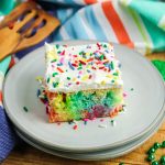 Rainbow Jello poke cake square topped with whipped topping and sprinkles on a plate.