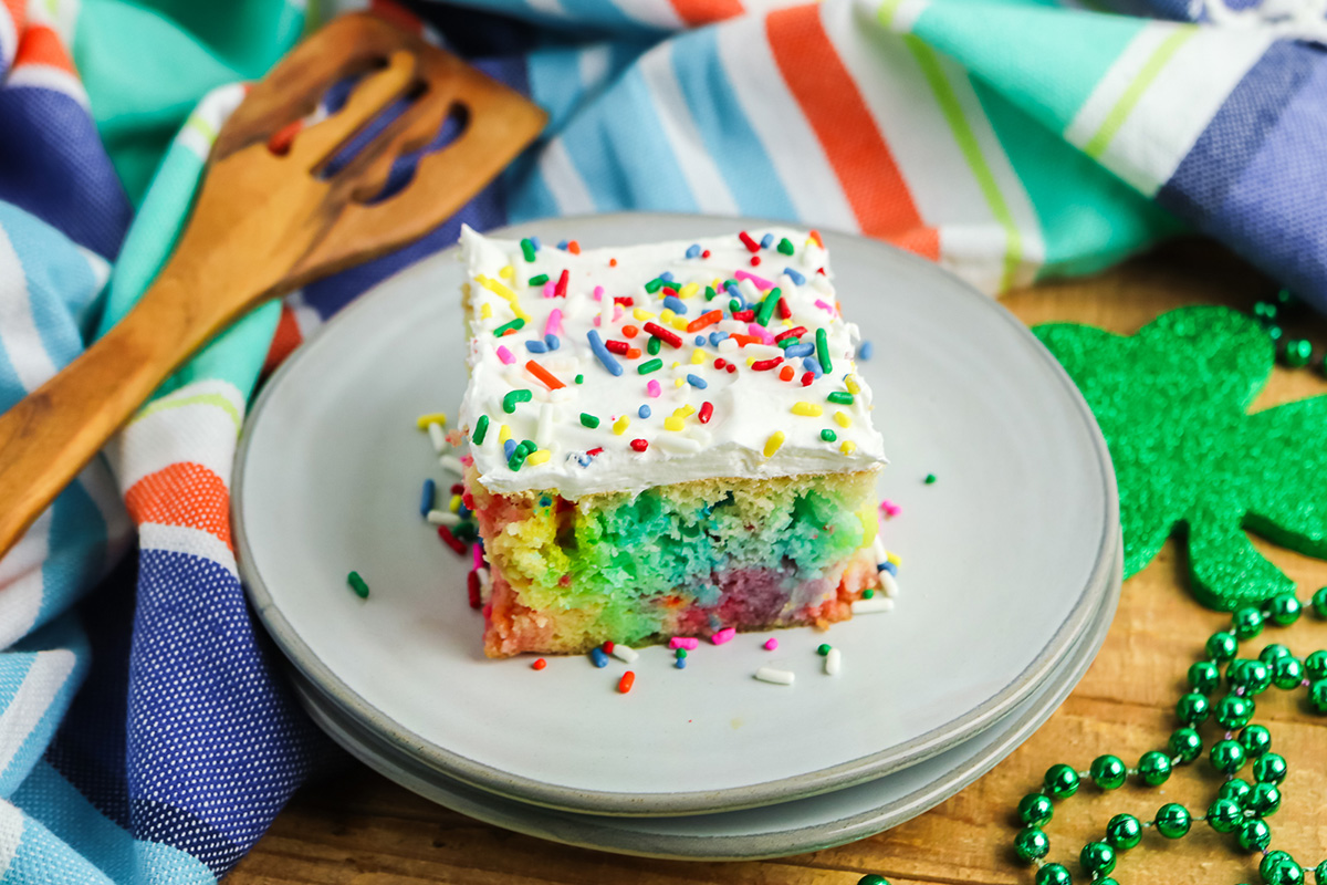 Rainbow Jello poke cake square topped with whipped topping and sprinkles on a plate.
