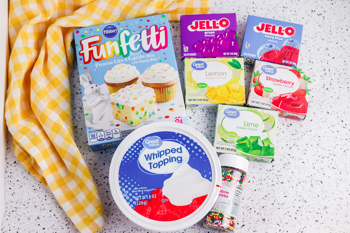 Ingredients for rainbow poke cake including Funfetti cake mix, Jello flavors, whipped topping, and sprinkles.