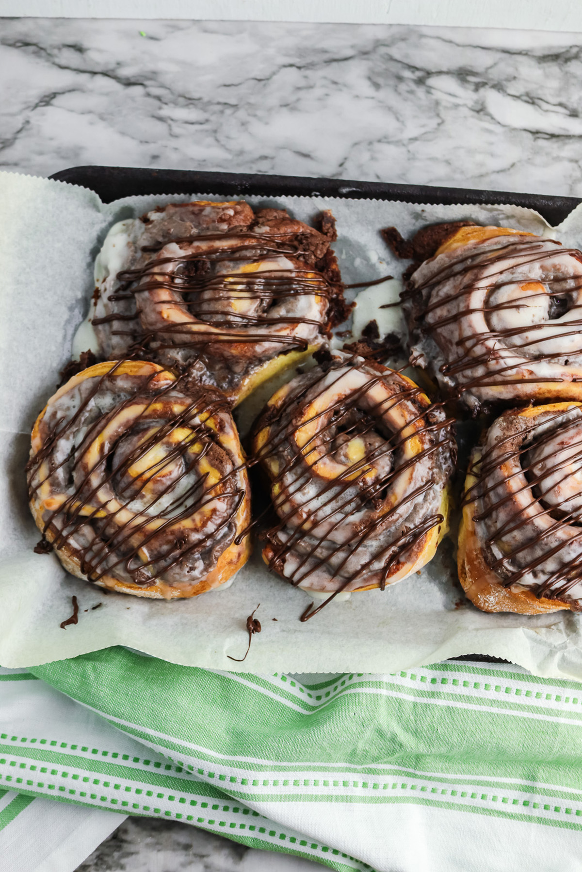 Brownie cinnamon rolls baked on parchment lined baking sheet with icing and melted chocolate drizzle on top.