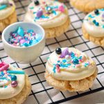 Cream cheese sugar cookies with frosting and colorful sprinkles cooling on a wire rack, with extra plain cookies on the side.