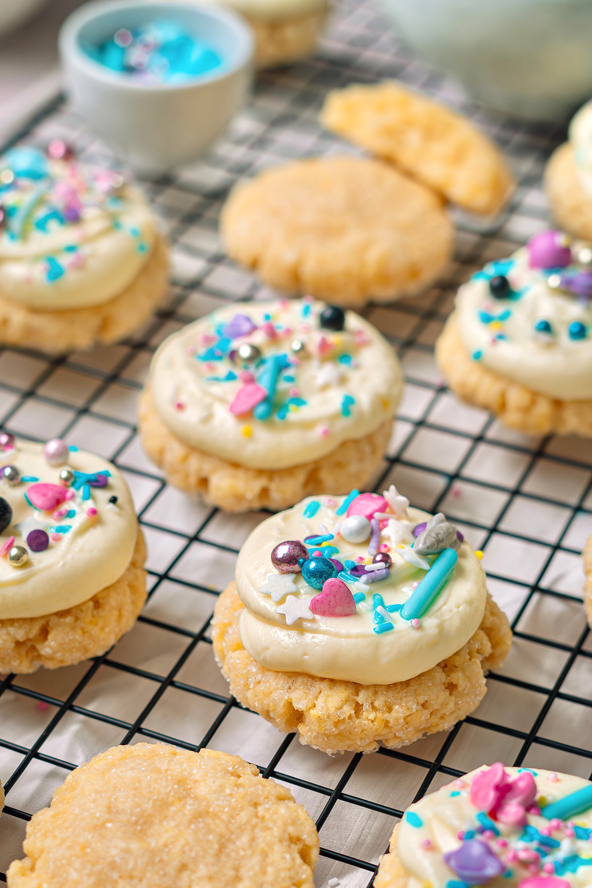 Close-up of cream cheese sugar cookies frosted with cream cheese icing and decorated with colorful sprinkles on a wire rack.