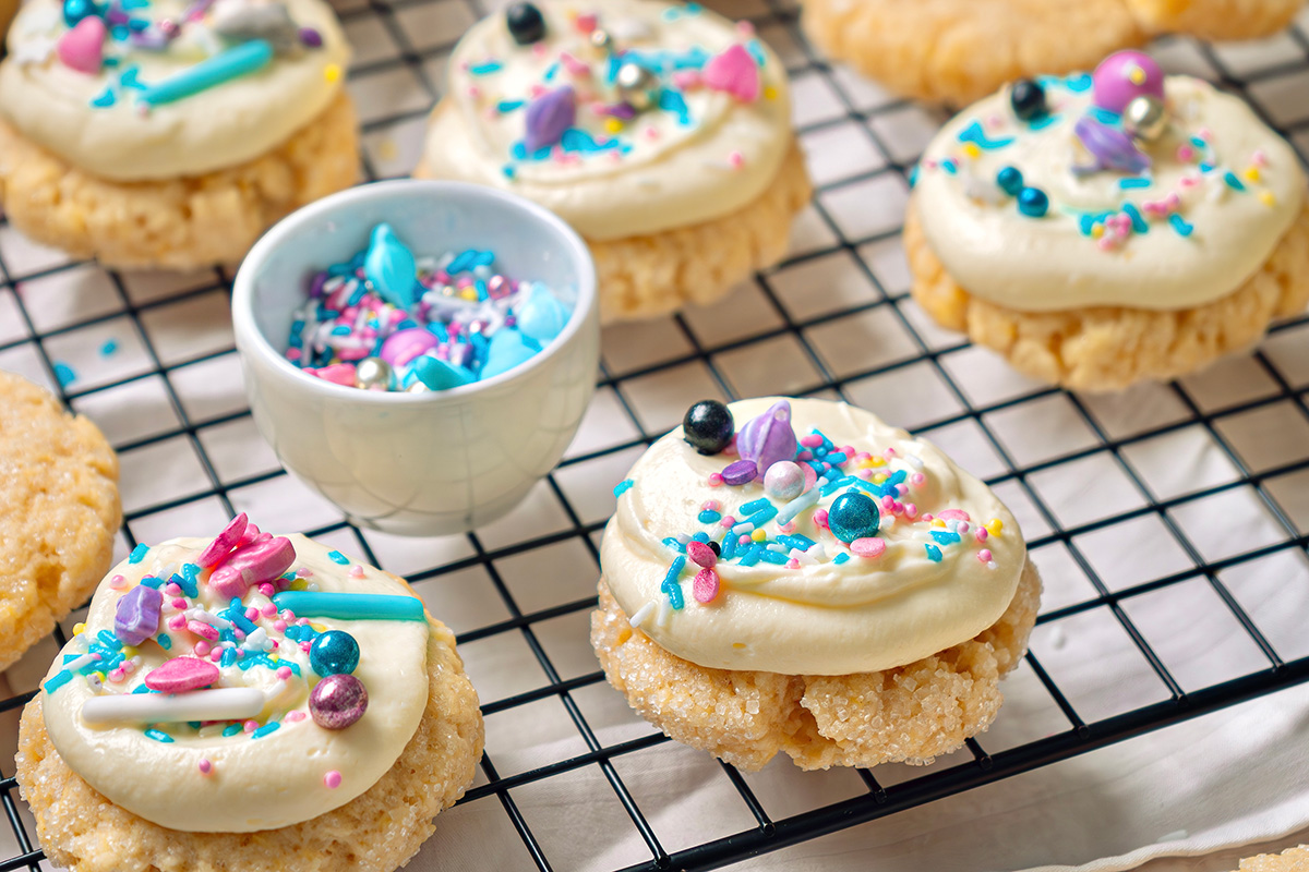 These cream cheese sugar cookies are soft, sweet, and topped with fluffy frosting. Starting with sugar cookie mix keeps them easy, but they still feel special enough for parties and holidays. Cream cheese sugar cookies with frosting and colorful sprinkles cooling on a wire rack, with extra plain cookies on the side.