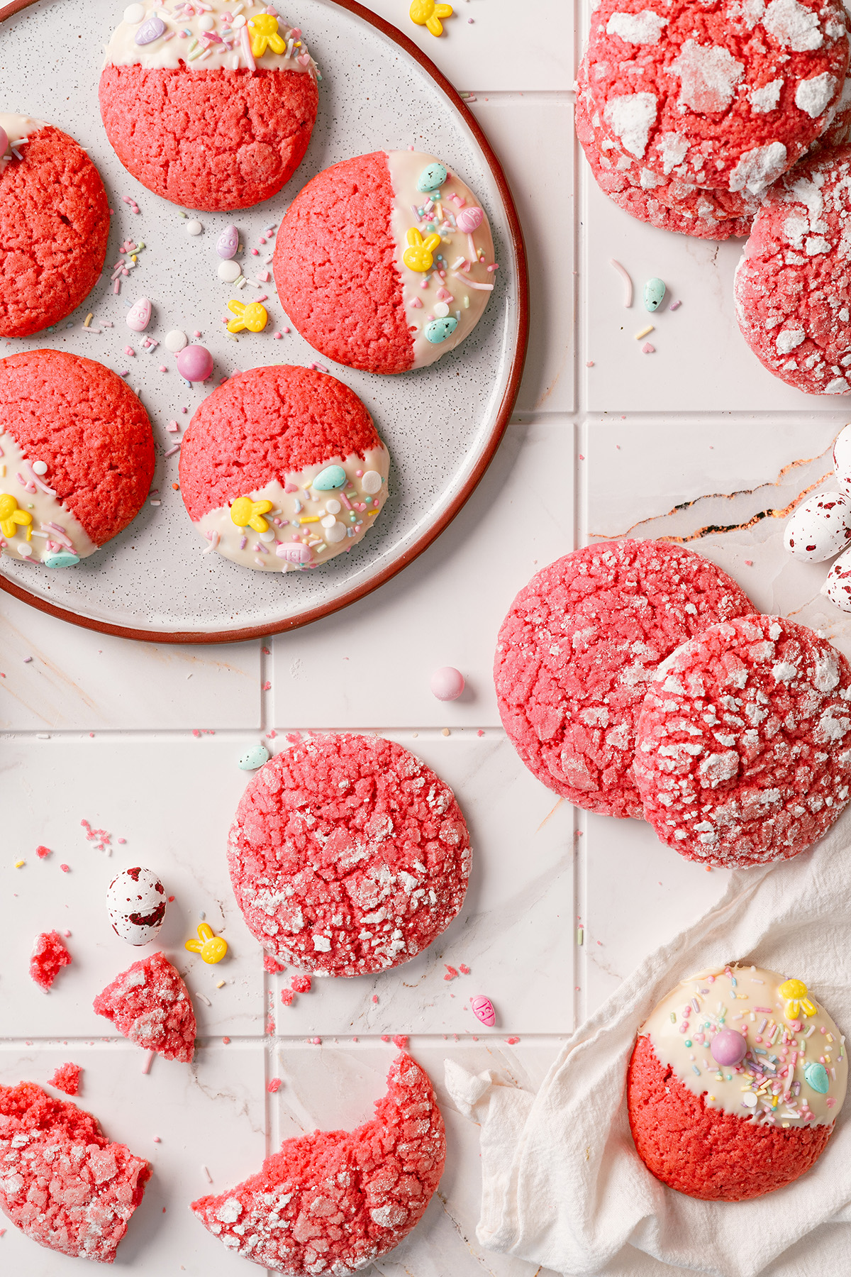 Pink Easter cake mix cookies on a white plate, some dipped in white chocolate and topped with pastel Easter sprinkles, with crinkle cookies and candy eggs scattered around.