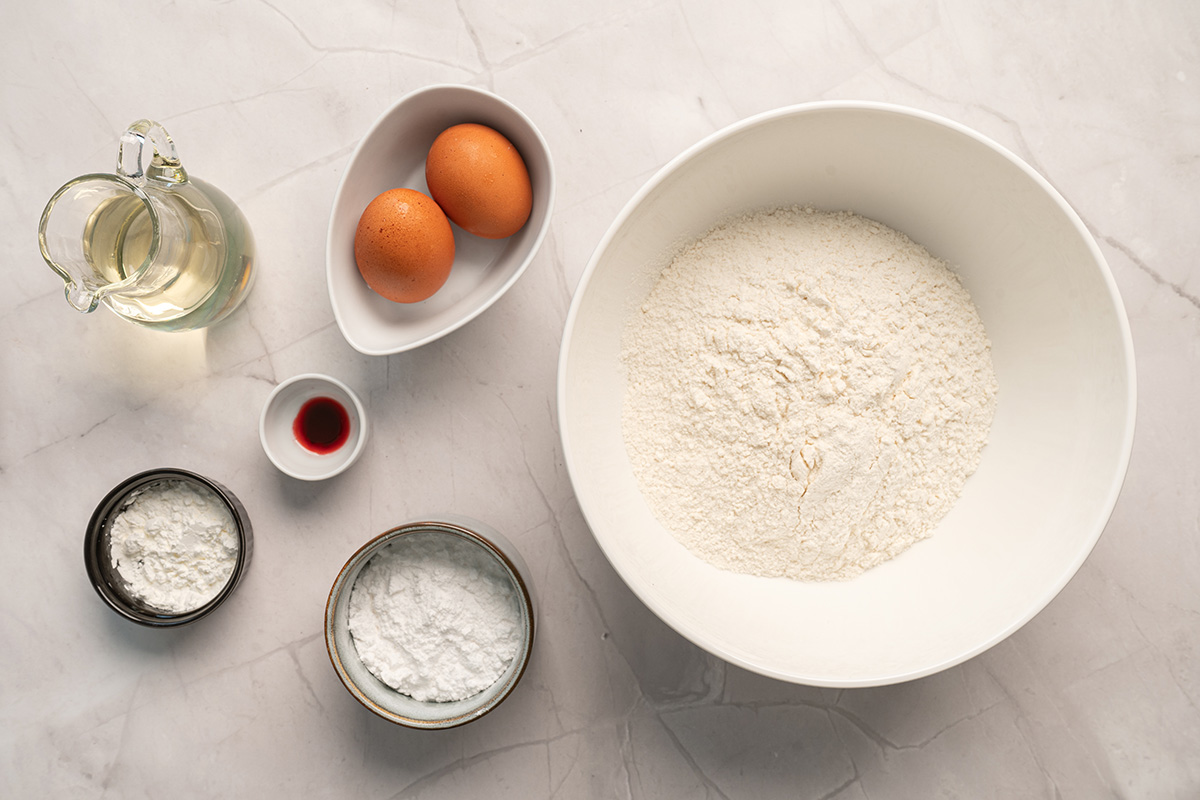 Ingredients for Easter cake mix cookies arranged on a marble surface, including white cake mix, eggs, oil, powdered sugar, cornstarch, and pink food coloring.