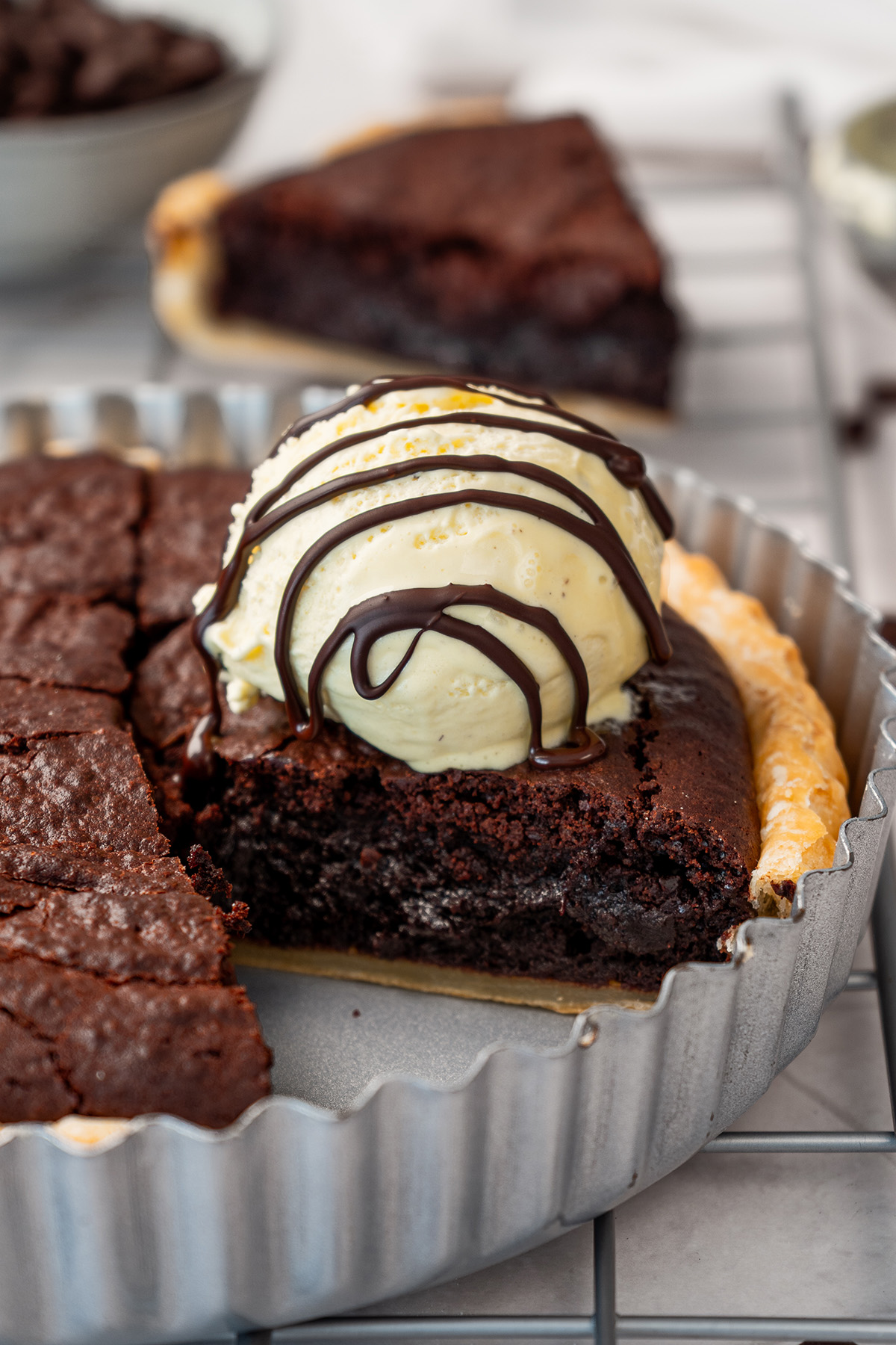 A brownie pie in its fluted tart pan on a cooling rack with a slice cut, topped with a scoop of vanilla ice cream and chocolate drizzle, with another plated slice visible in the background.