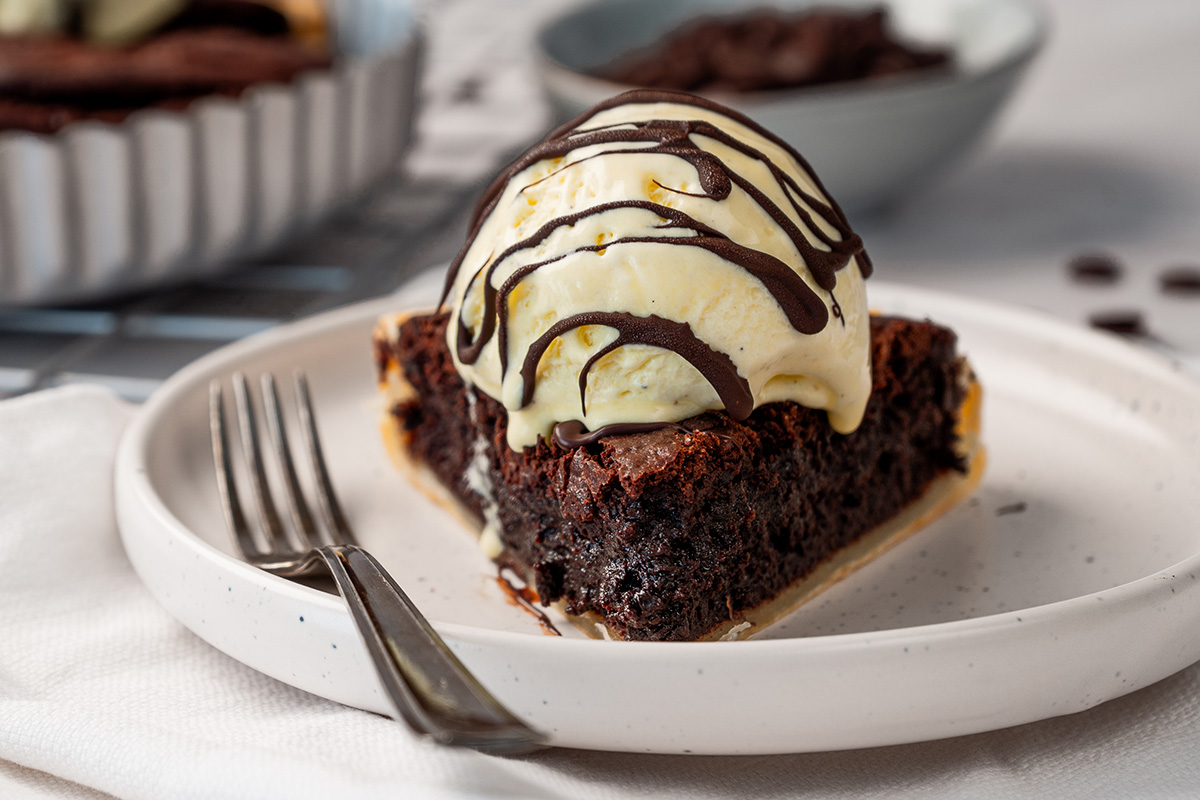 A slice of fudgy brownie pie with a flaky crust, topped with a scoop of vanilla ice cream and chocolate drizzle, on a speckled plate with the whole pie visible in the background.