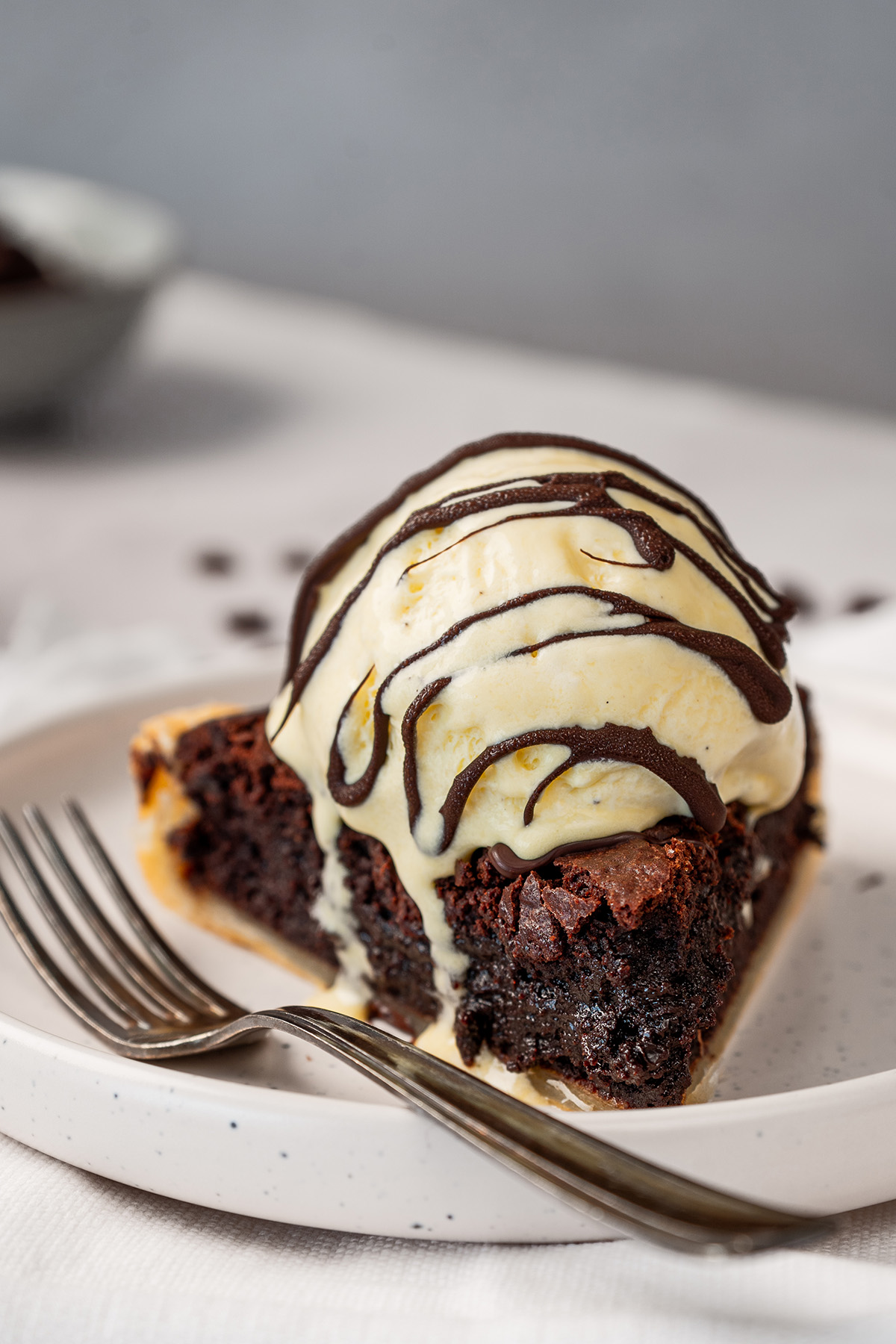 A close-up of a slice of fudgy brownie pie topped with a scoop of vanilla ice cream drizzled with chocolate sauce, served on a speckled white plate with a fork.