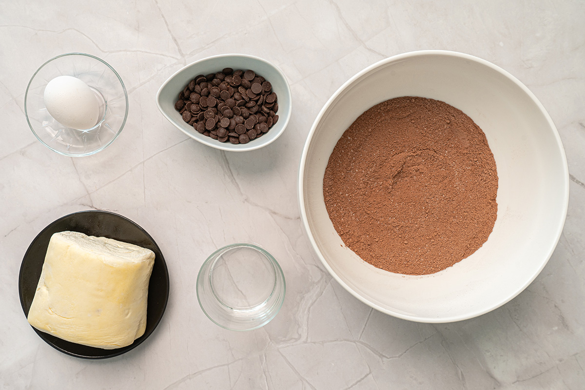 Overhead shot of brownie pie ingredients laid out: a bowl of brownie mix, a small bowl of semi-sweet chocolate chips, one egg in a glass dish, a block of refrigerated pie crust dough, and a small glass of water.
