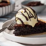 A close-up angled shot of a brownie pie slice topped with melting vanilla ice cream and chocolate sauce, showing the dense, fudgy interior texture and buttery crust.