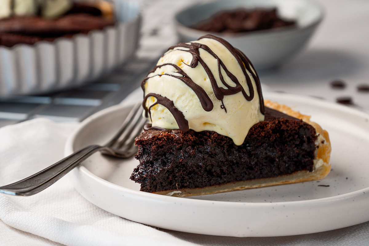 A close-up angled shot of a brownie pie slice topped with melting vanilla ice cream and chocolate sauce, showing the dense, fudgy interior texture and buttery crust.