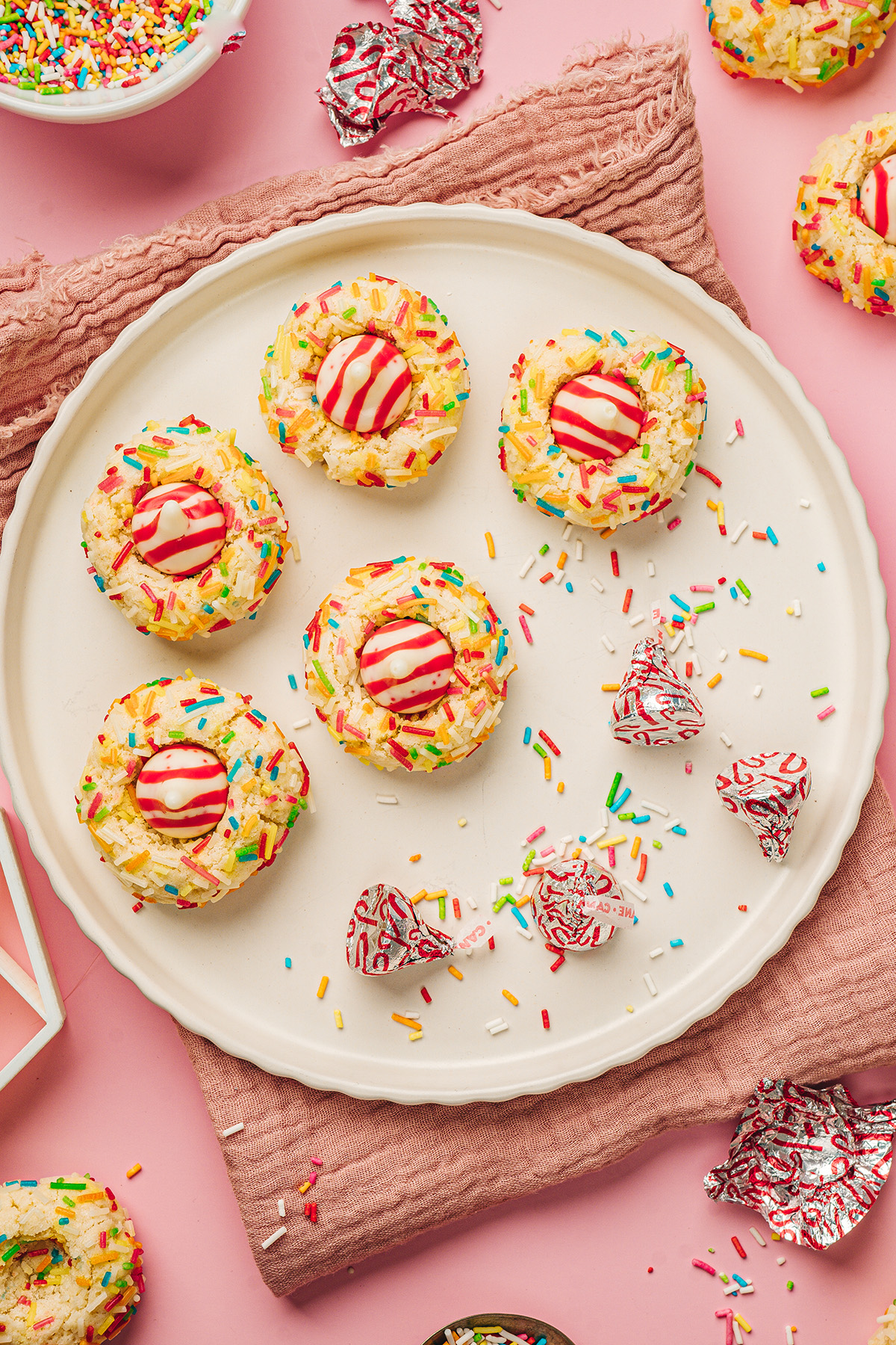 Sugar cookie blossoms topped with Hershey’s Hugs and rainbow sprinkles served on a cream plate over a pink background.