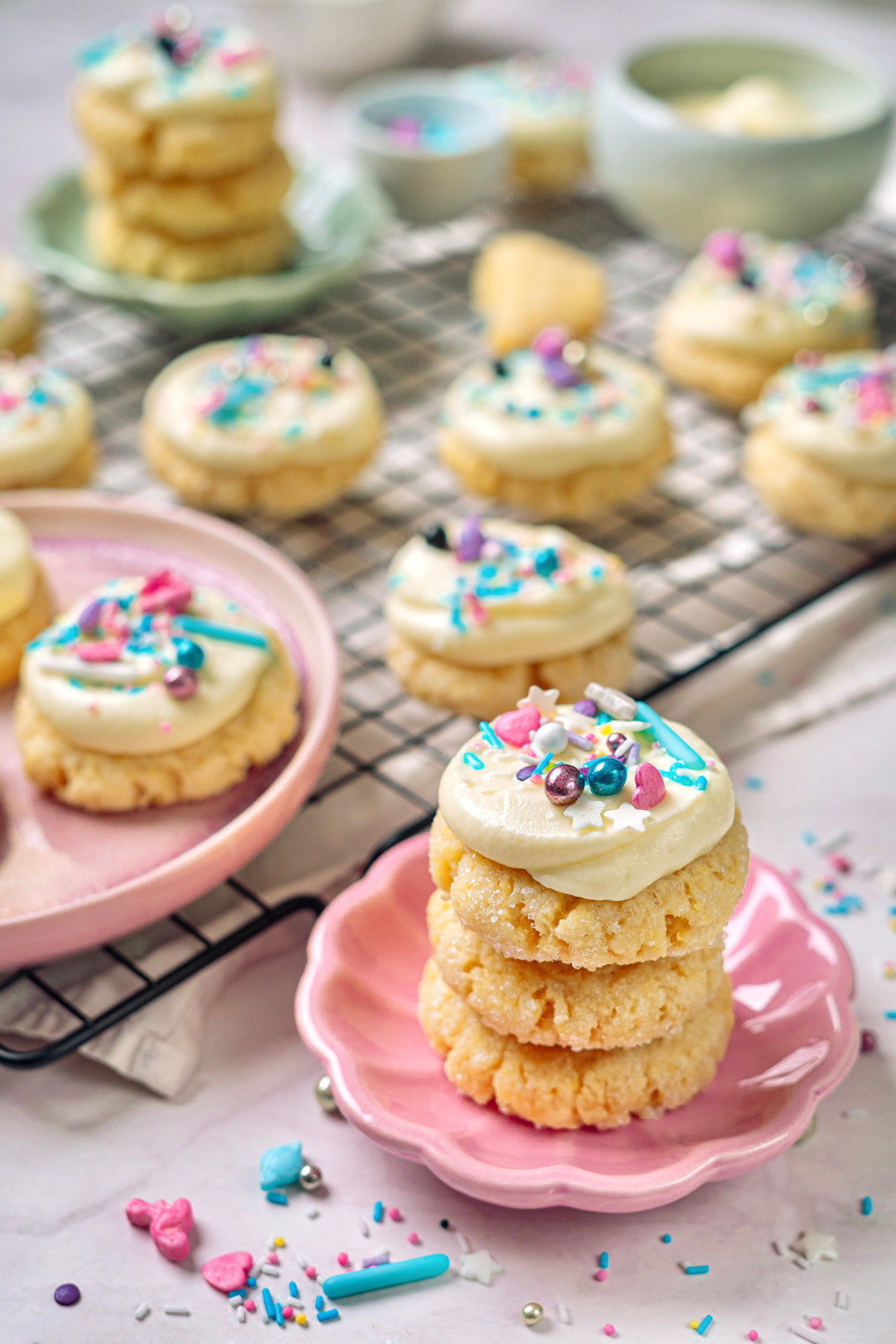 These cream cheese sugar cookies are soft, sweet, and topped with fluffy frosting. Starting with sugar cookie mix keeps them easy, but they still feel special enough for parties and holidays. Stack of frosted cream cheese sugar cookies with pastel sprinkles on a pink plate, with more cookies in the background.