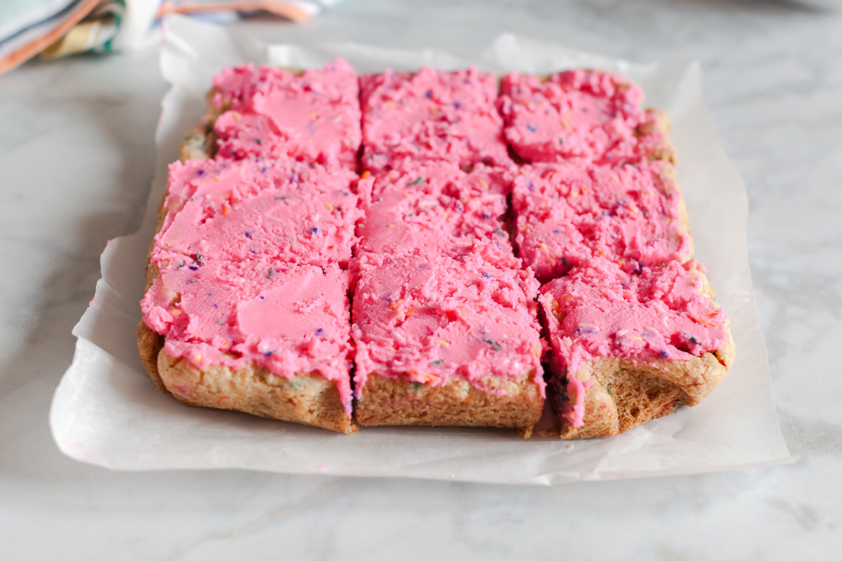These funfetti sugar cookie bars start with a shortcut and bake up soft, buttery, and packed with sprinkles. Finished with a thick layer of pink frosting, they’re the kind of treat that disappears fast. Funfetti sugar cookie bars cut into squares with pink frosting spread evenly on top.