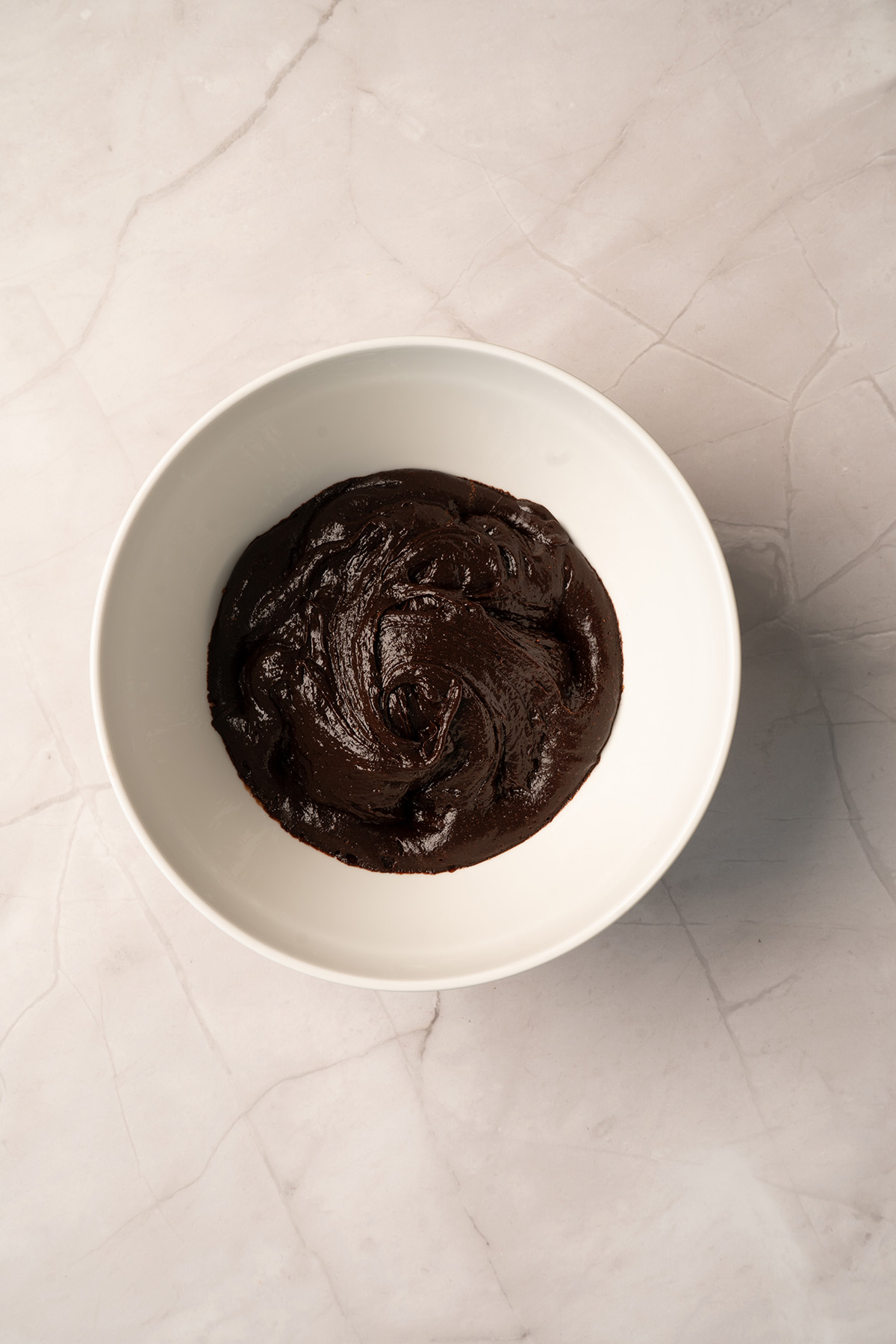 A white bowl filled with thick, dark chocolate brownie batter stirred together, viewed from above on a marble surface.