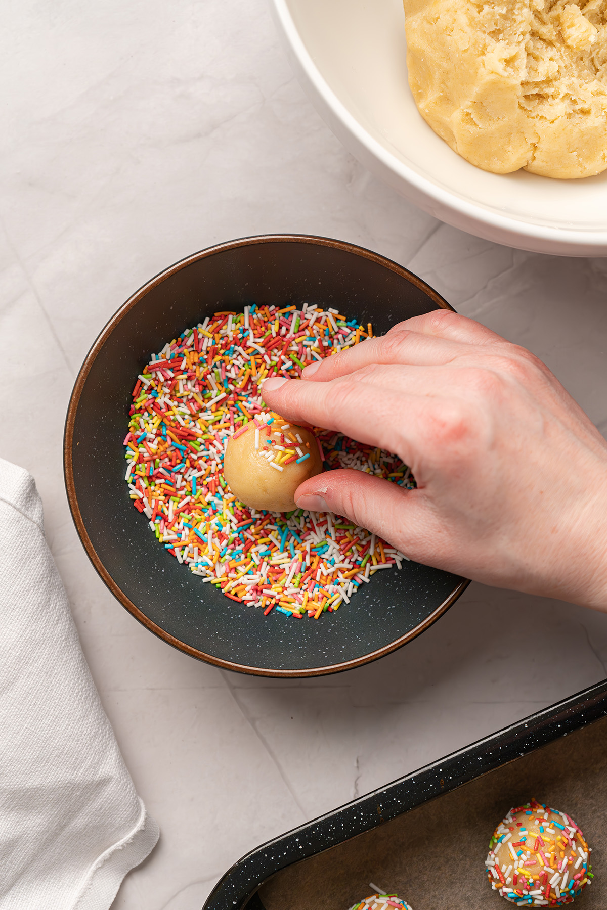 Hand rolling a sugar cookie dough ball in rainbow sprinkles before baking, with dough bowl nearby.