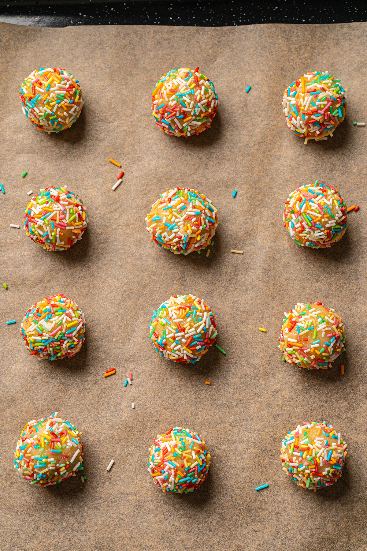 Rainbow sprinkle-coated sugar cookie dough balls arranged on a parchment-lined baking sheet before baking.