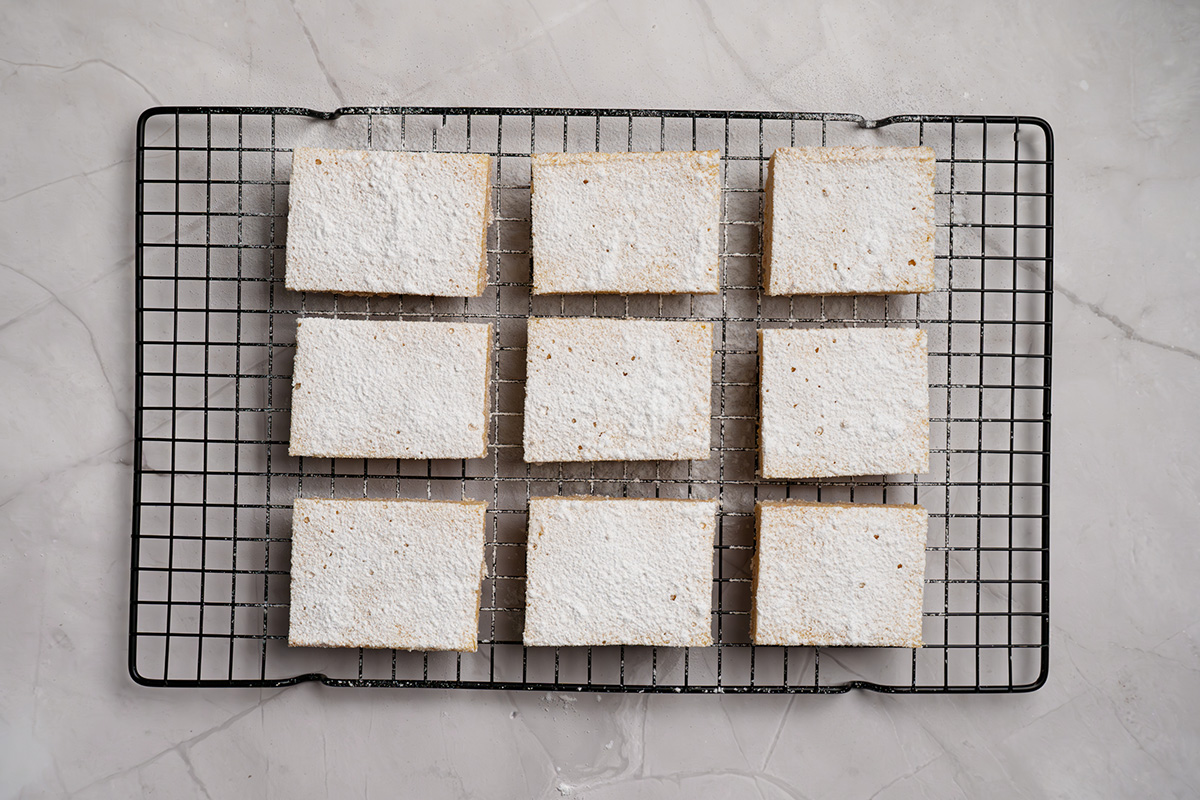 Easy sugar cookie lemon bars made with cookie mix crust, lemon juice, and zest. A sweet, tangy treat to chill and slice. Sugar cookie lemon bars cut into squares on a cooling rack with powdered sugar.