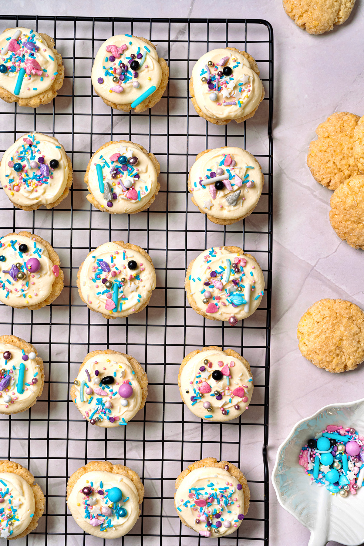These cream cheese sugar cookies are soft, sweet, and topped with fluffy frosting. Starting with sugar cookie mix keeps them easy, but they still feel special enough for parties and holidays. Cream cheese sugar cookies with frosting and colorful sprinkles cooling on a wire rack, with extra plain cookies on the side.