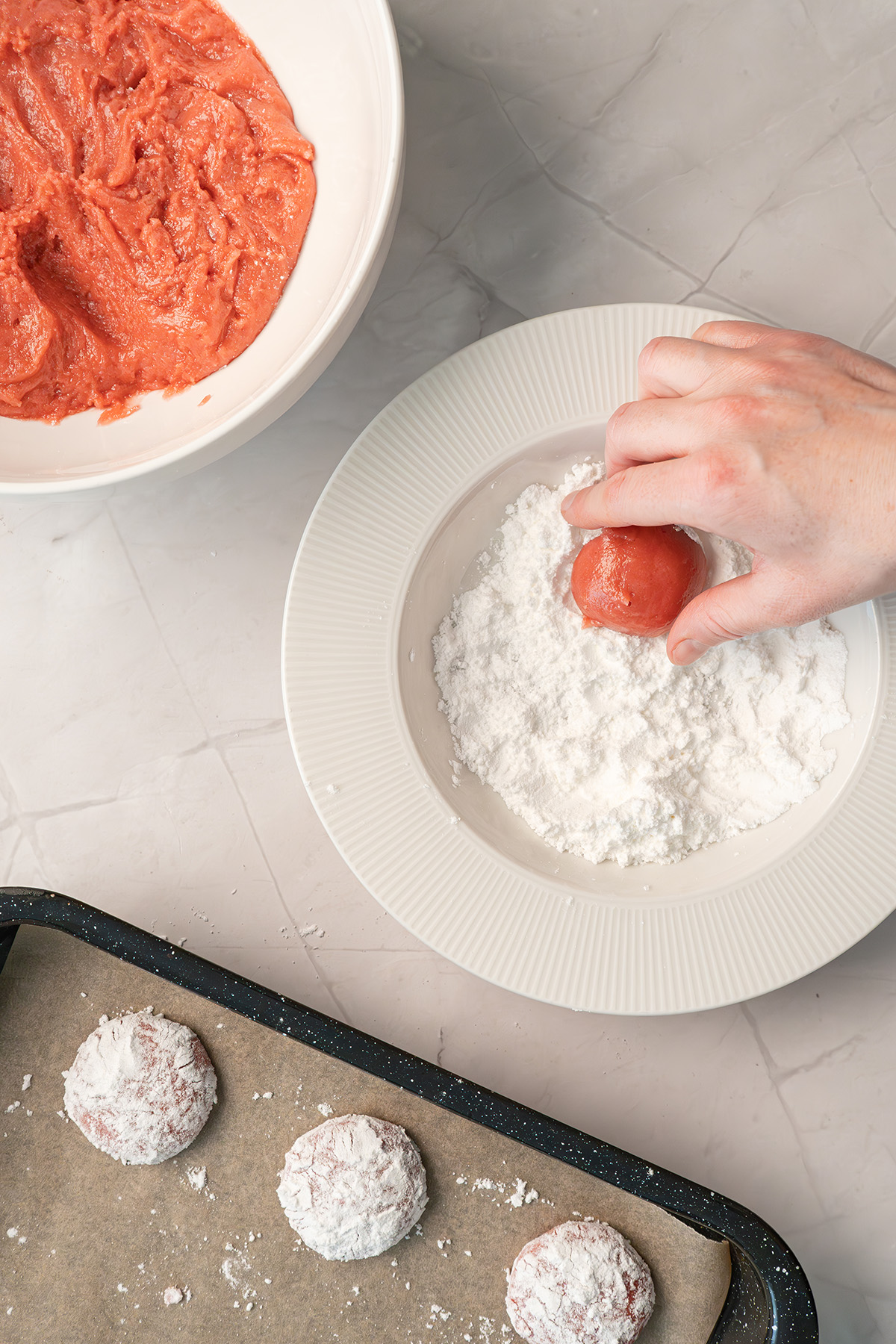 Hand rolling pink cookie dough in powdered sugar beside a baking sheet of coated cookie dough balls.