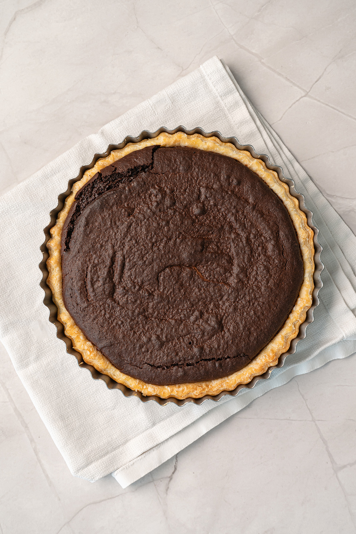 A fully baked fudgy brownie pie in a fluted tart pan, with a deep chocolate filling and golden baked crust, resting on a white linen napkin on a marble surface.