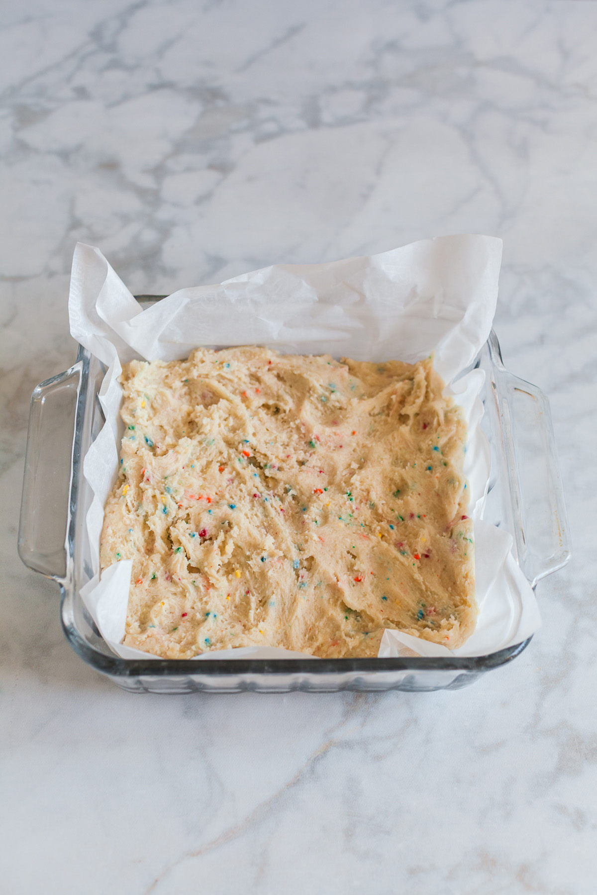 These funfetti sugar cookie bars start with a shortcut and bake up soft, buttery, and packed with sprinkles. Finished with a thick layer of pink frosting, they’re the kind of treat that disappears fast. Funfetti sugar cookie dough pressed evenly into a parchment-lined square baking dish before baking.