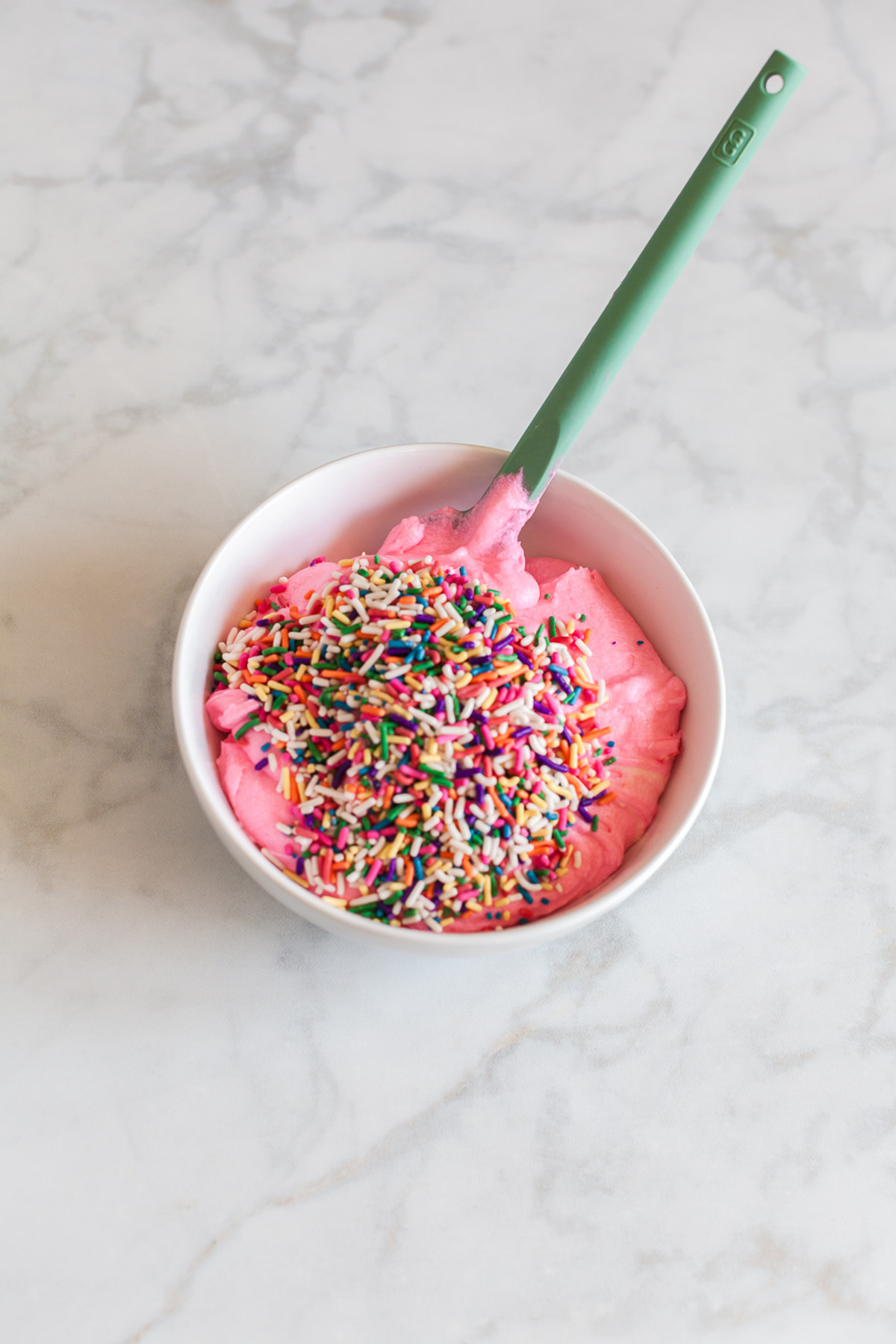 These funfetti sugar cookie bars start with a shortcut and bake up soft, buttery, and packed with sprinkles. Finished with a thick layer of pink frosting, they’re the kind of treat that disappears fast. Pink buttercream frosting in a bowl topped with rainbow sprinkles and a spatula.
