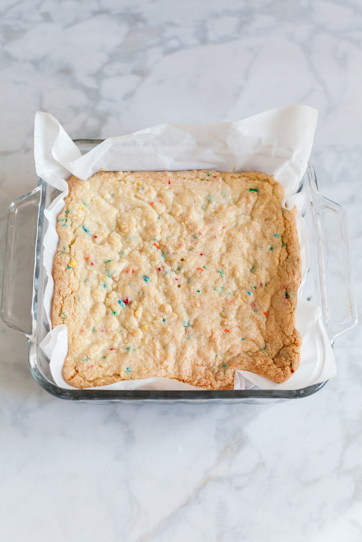 These funfetti sugar cookie bars start with a shortcut and bake up soft, buttery, and packed with sprinkles. Finished with a thick layer of pink frosting, they’re the kind of treat that disappears fast. Baked funfetti sugar cookie bar base in a parchment-lined glass baking dish with lightly golden edges.