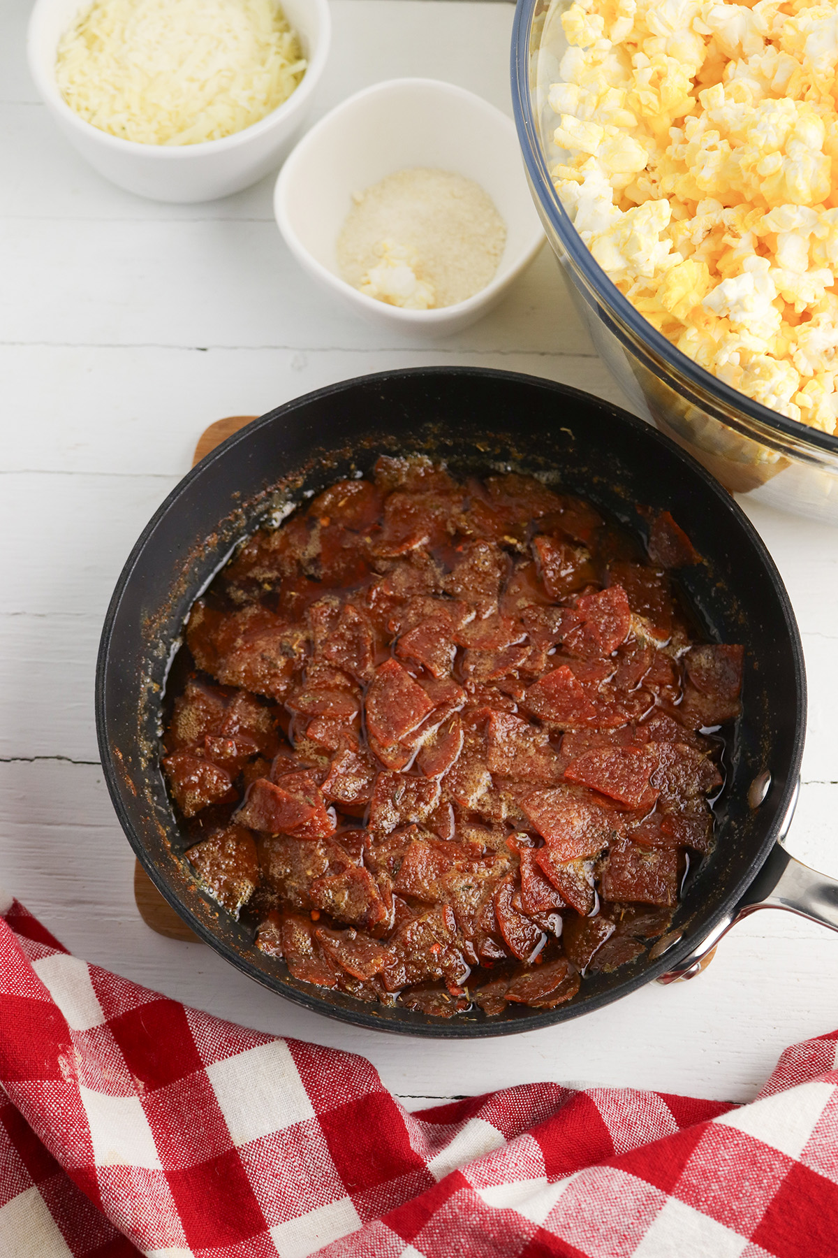 A skillet filled with chopped pepperoni sautéed in melted butter with Italian seasoning and spices, with bowls of shredded cheese, parmesan, and a glass bowl of popcorn in the background.