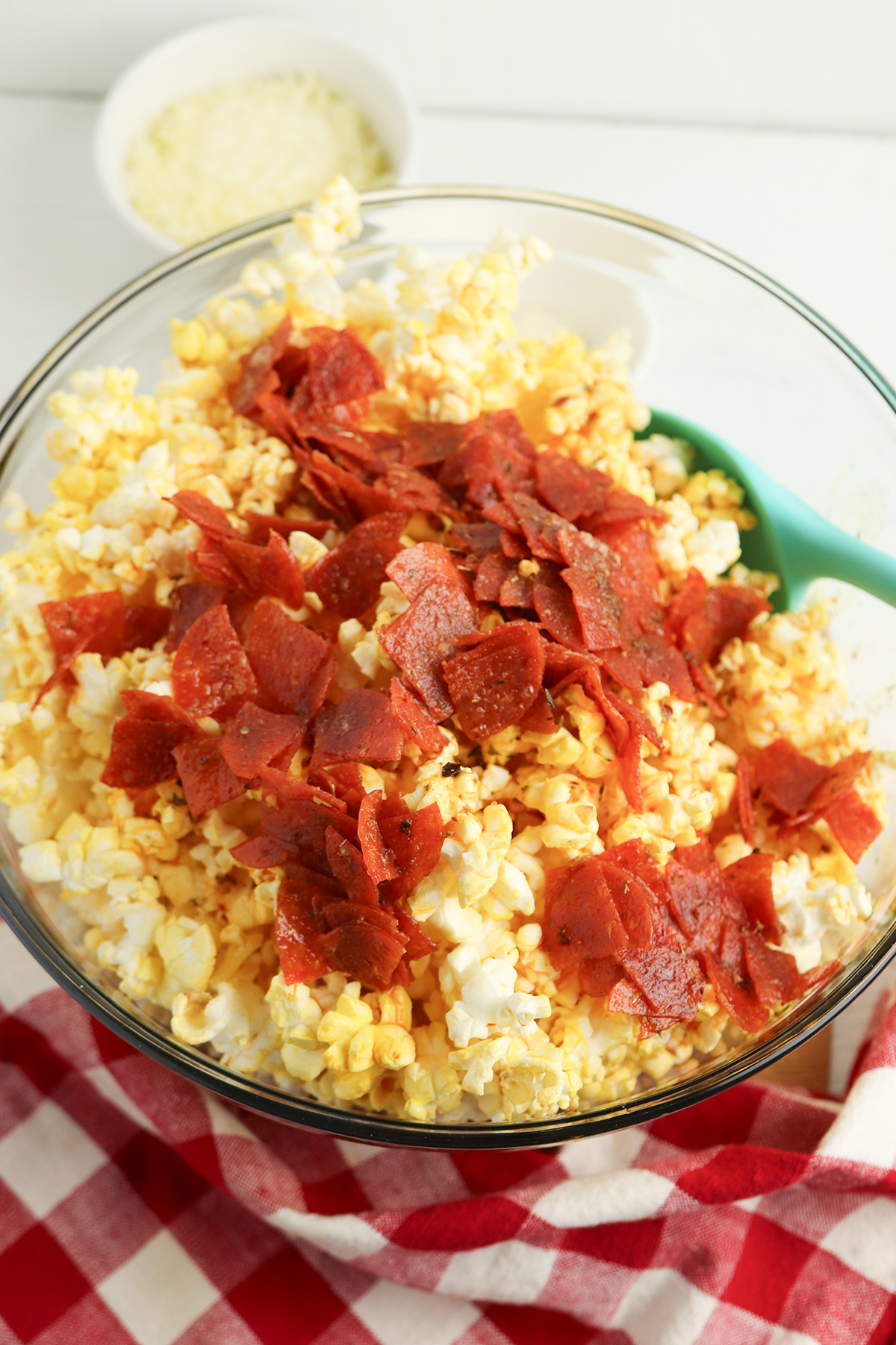 The seasoned butter and pepperoni mixture poured over popcorn in a large glass bowl, with a teal spatula.
