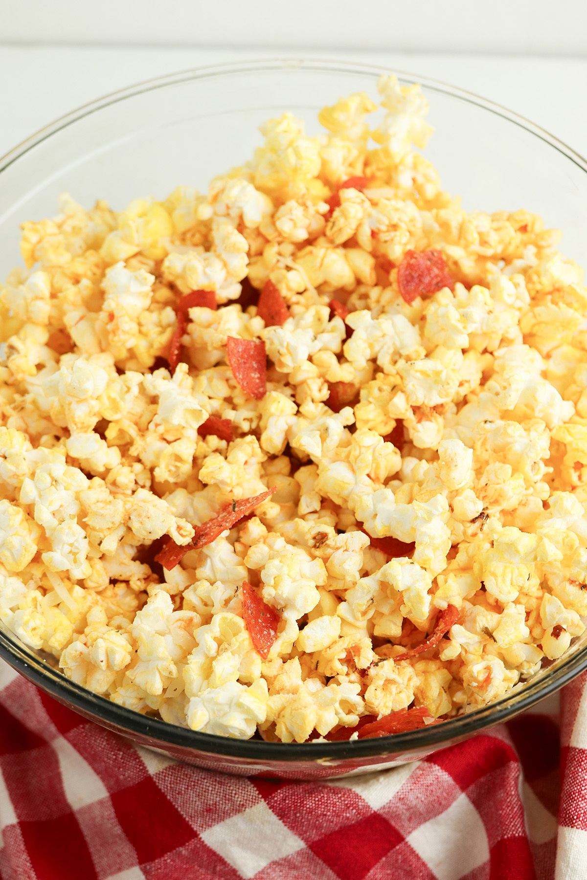 The fully tossed pepperoni pizza popcorn in a glass bowl on a red gingham cloth, with the cheese melted and coating the popcorn evenly alongside the pepperoni pieces.