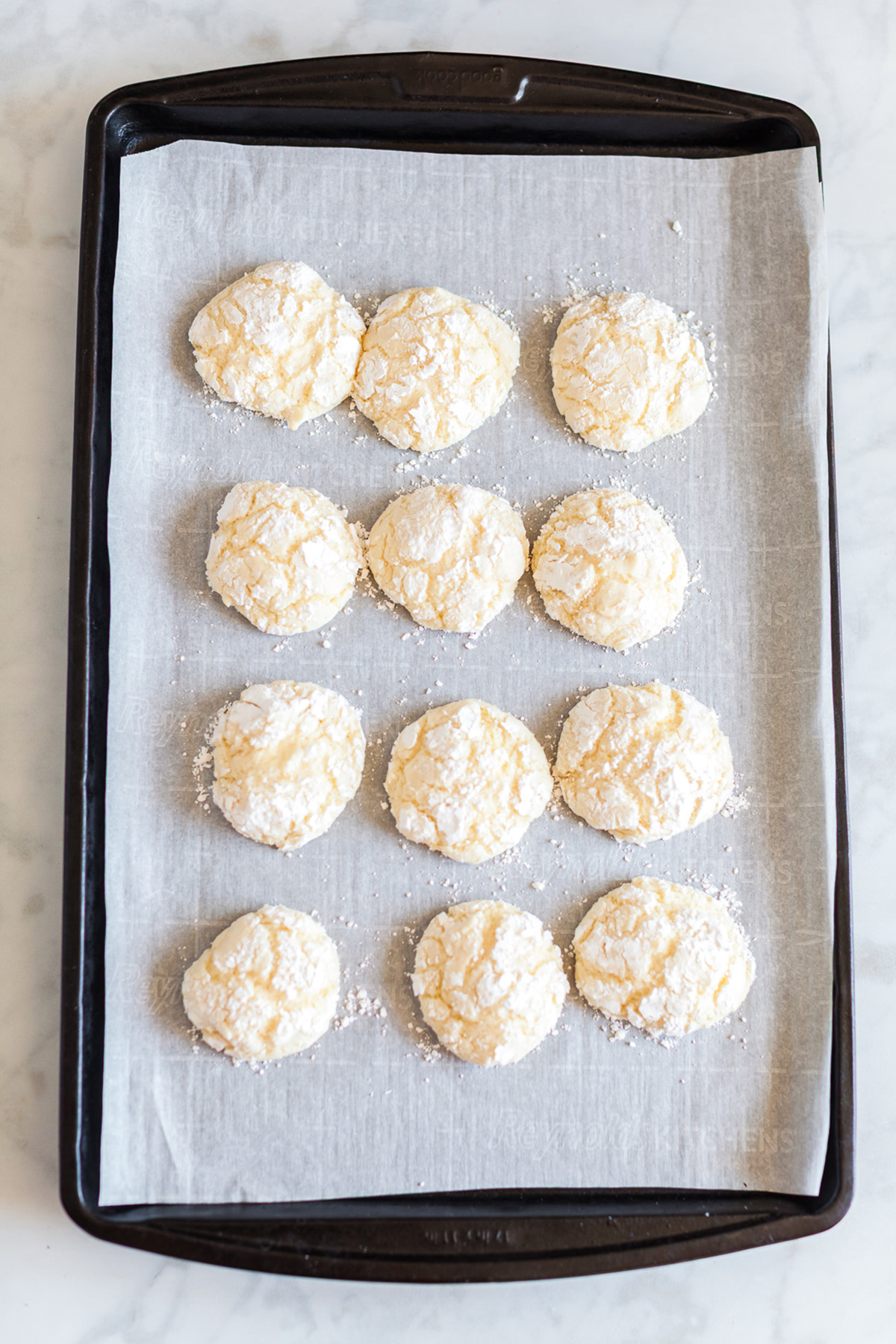 Baked vanilla crinkle cookie dough balls coated in powdered sugar on a parchment lined baking sheet.