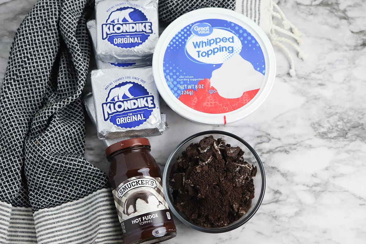 Klondike ice cream cake ingredients including Klondike bars, whipped topping, hot fudge, and crushed Oreo cookies on a marble surface.