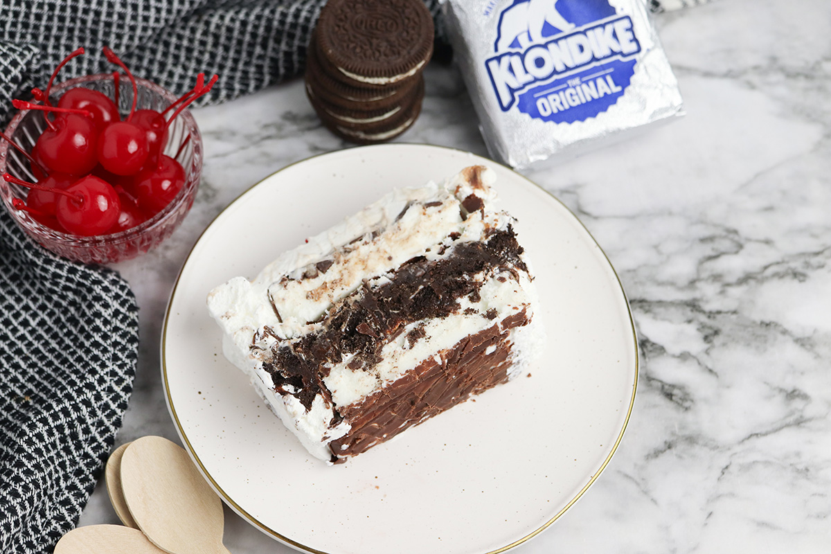 Klondike ice cream cake slice on a plate with cherries, Oreo cookies, and Klondike bars in the background.