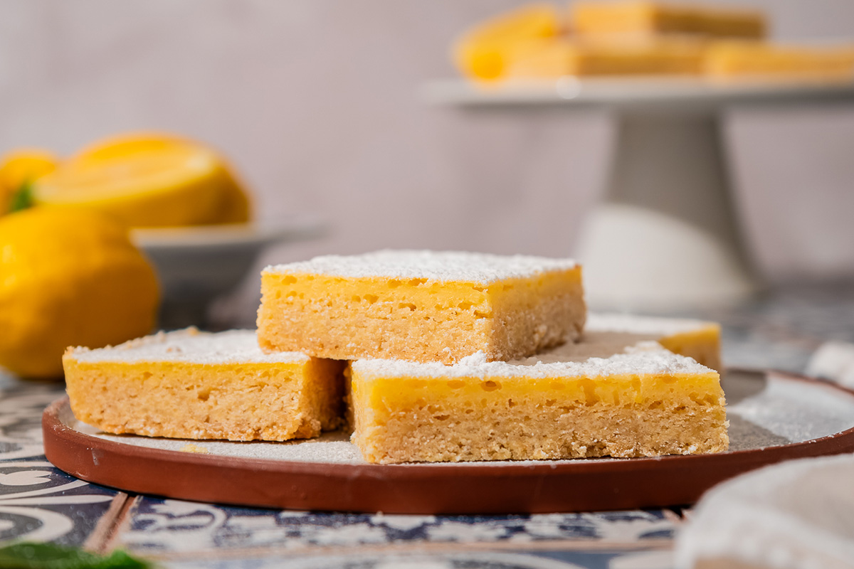 Easy sugar cookie lemon bars made with cookie mix crust, lemon juice, and zest. A sweet, tangy treat to chill and slice. Sugar cookie lemon bars stacked on a plate, dusted with powdered sugar.