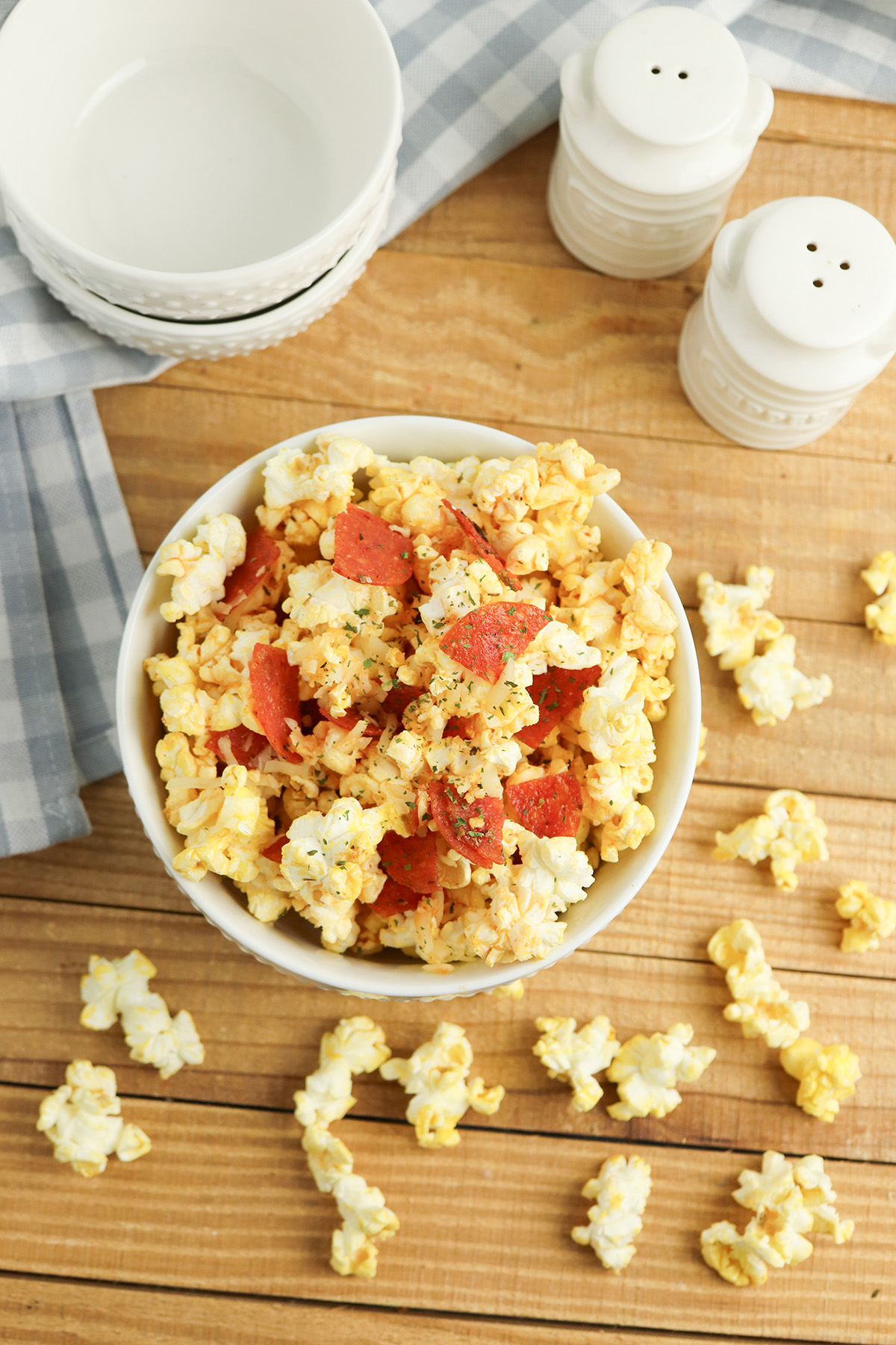Overhead view of a white bowl of pepperoni pizza popcorn on a wooden table, surrounded by salt and pepper shakers, stacked white bowls, a blue gingham napkin, and scattered popcorn pieces.