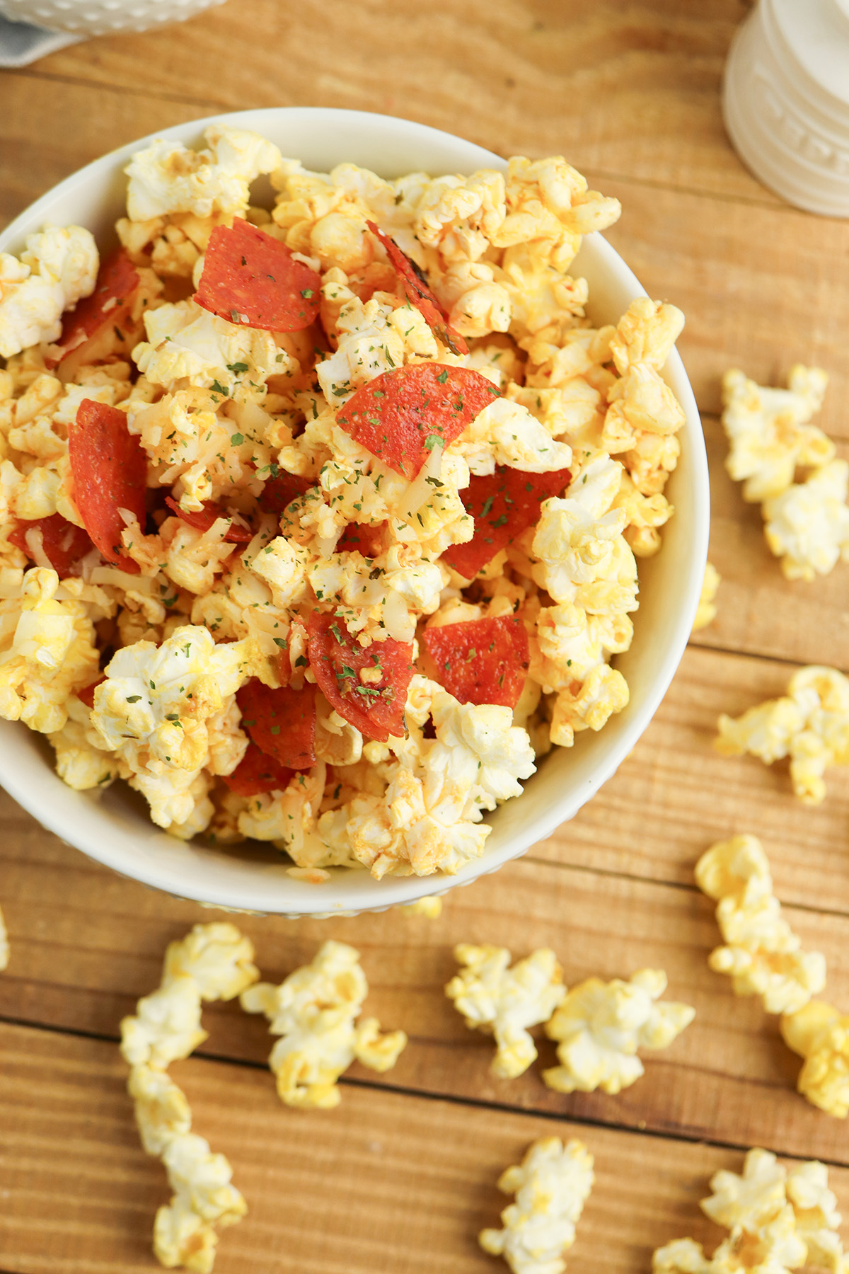 A close-up overhead view of a white bowl filled with pepperoni pizza popcorn, showing the pepperoni chunks, herbs, and cheese coating in detail, with scattered popcorn kernels on the wooden table around it.