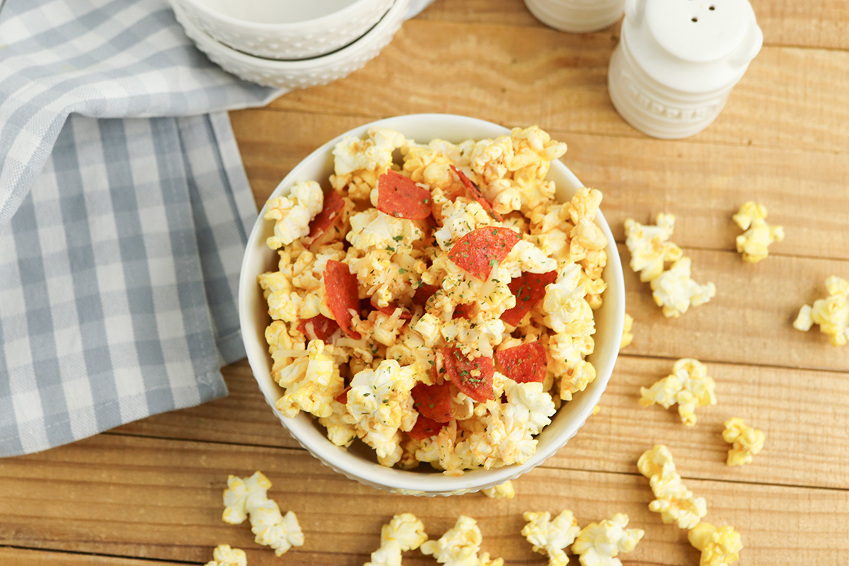 A large glass bowl filled with finished pepperoni pizza popcorn, showing buttery popcorn tossed with chopped pepperoni pieces and melted cheese, sitting on a red and white gingham cloth.