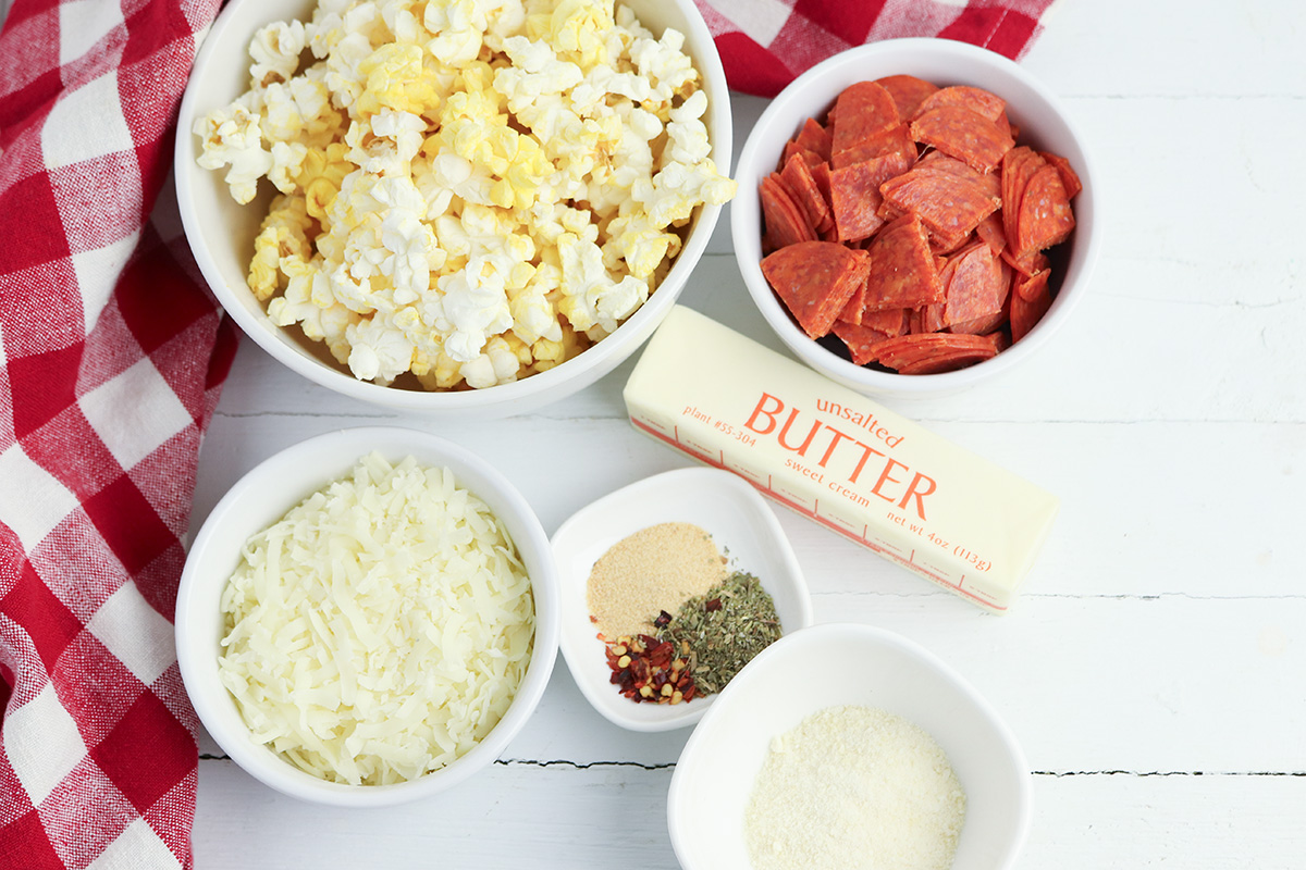 Flat lay of all recipe ingredients including popped popcorn, chopped pepperoni, shredded pizza blend cheese, grated parmesan, a small bowl of Italian seasoning with garlic powder and red pepper flakes, and a stick of unsalted butter.