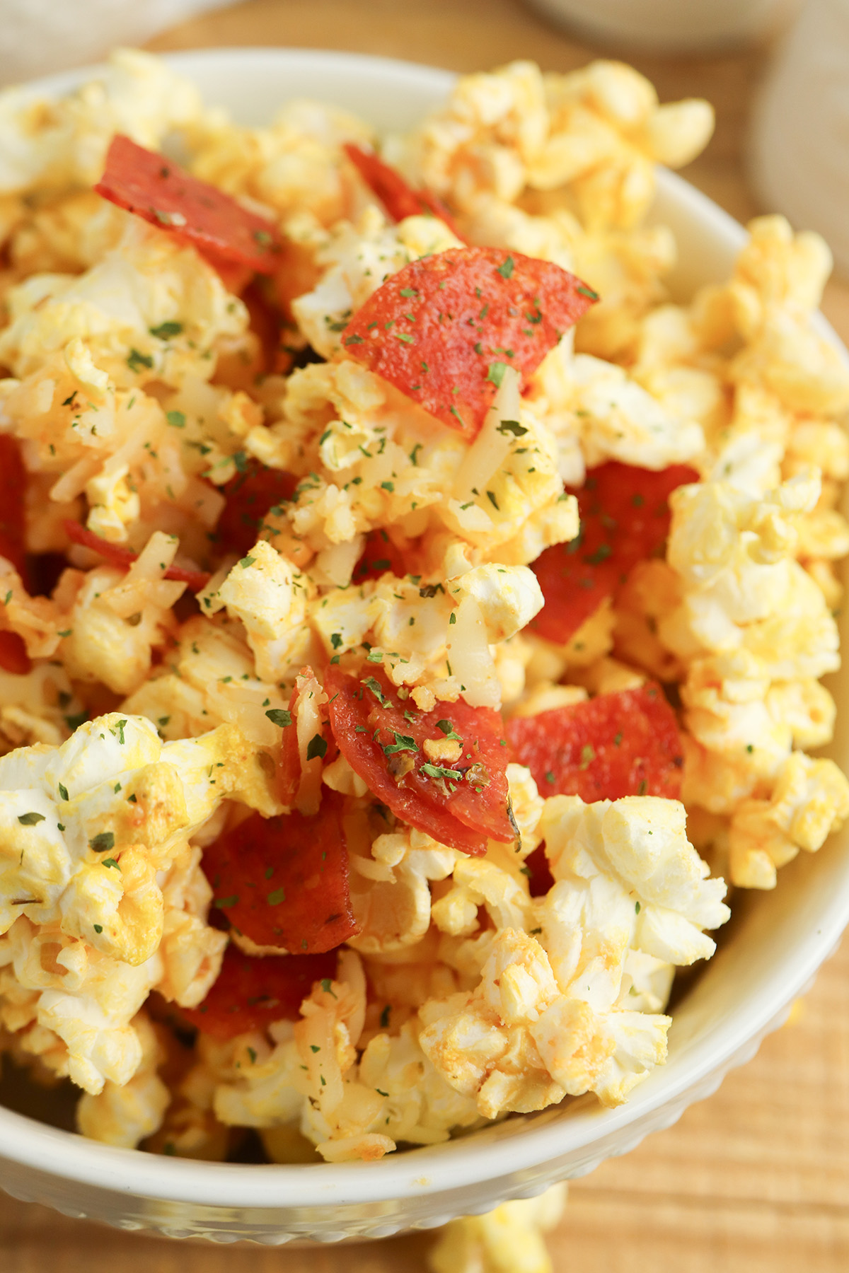 An angled close-up of pepperoni popcorn in a white pedestal bowl, highlighting the chopped pepperoni, melted cheese, and green herb flecks on the popcorn.
