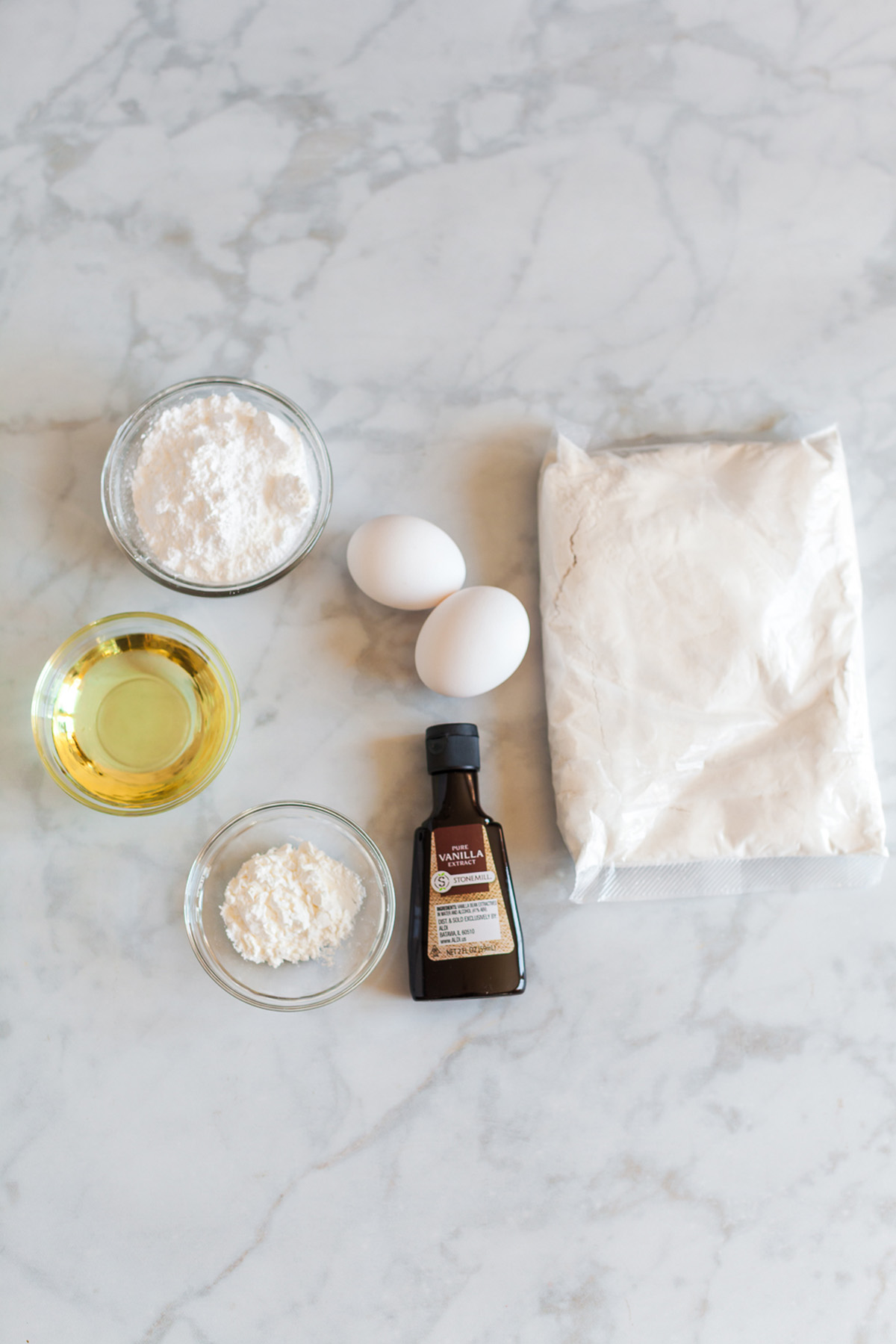 Vanilla cake mix crinkle cookie ingredients including white cake mix, eggs, vegetable oil, powdered sugar, cornstarch, and vanilla extract on a marble surface.
