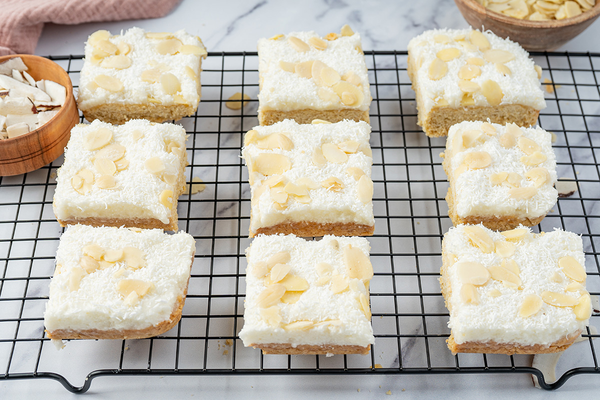 Frosted coconut sugar cookie bars topped with shredded coconut and sliced almonds on a cooling rack.