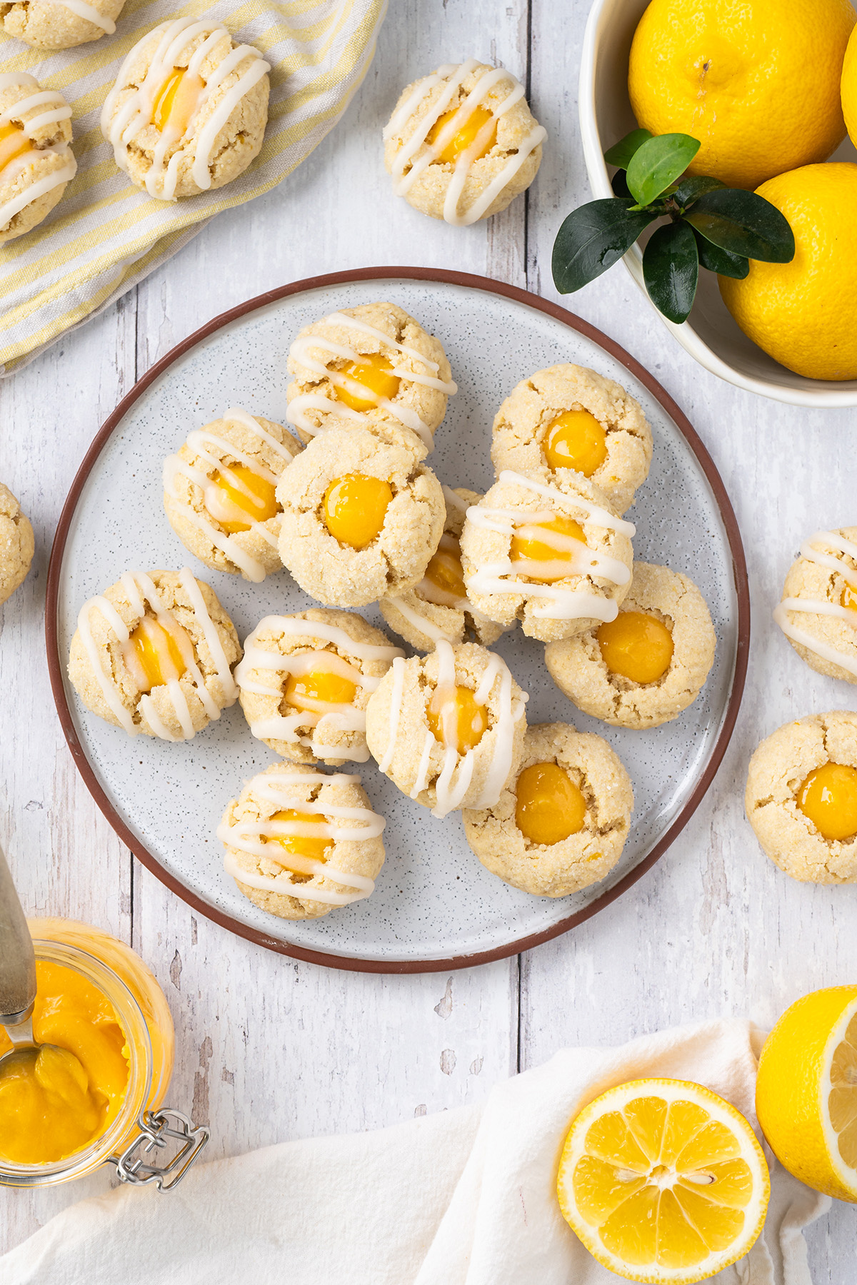 Lemon thumbprint cookies arranged on a plate with icing drizzle, surrounded by fresh lemons and lemon curd.