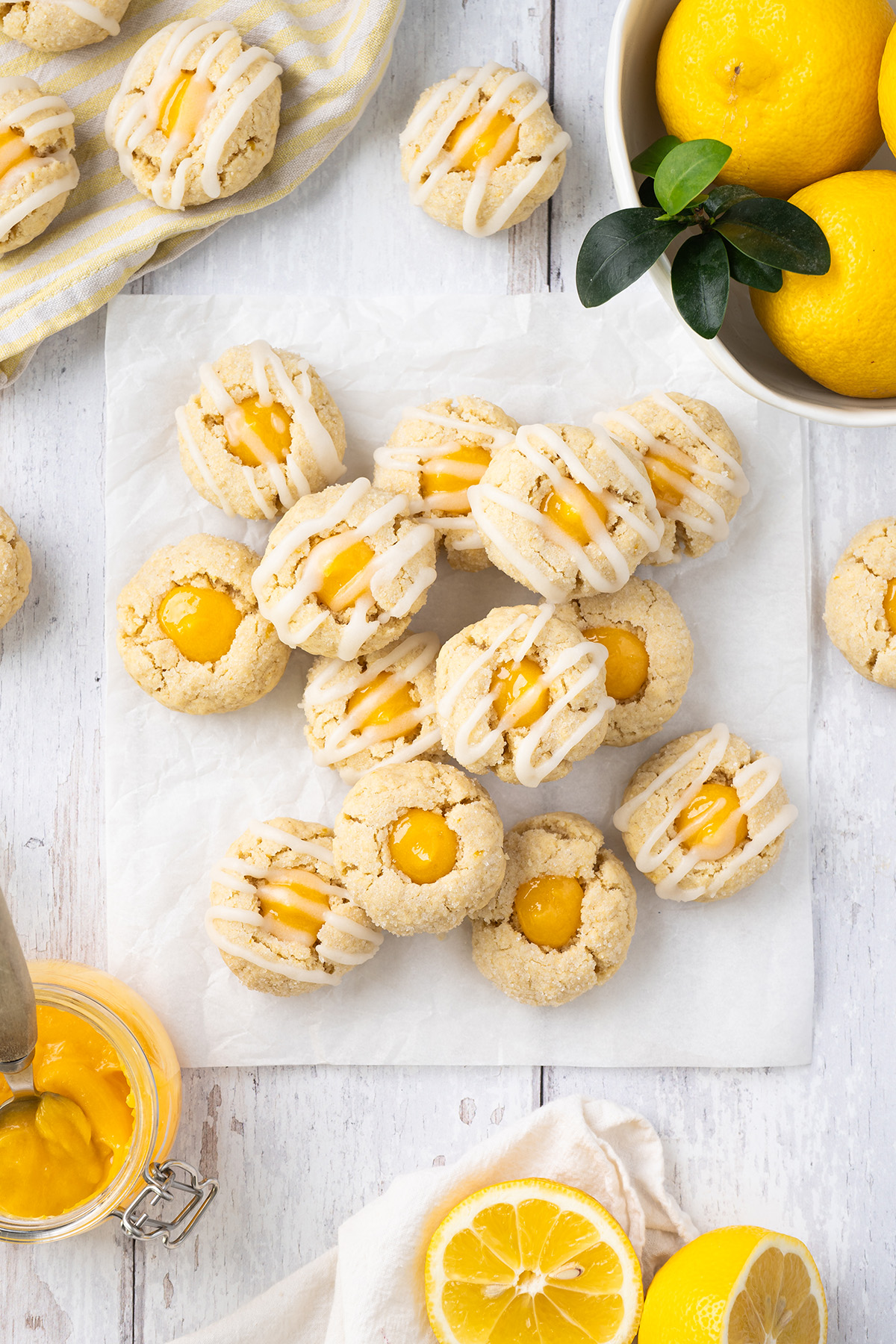 Lemon thumbprint cookies drizzled with icing and filled with lemon curd, styled with fresh lemons and a jar of curd.