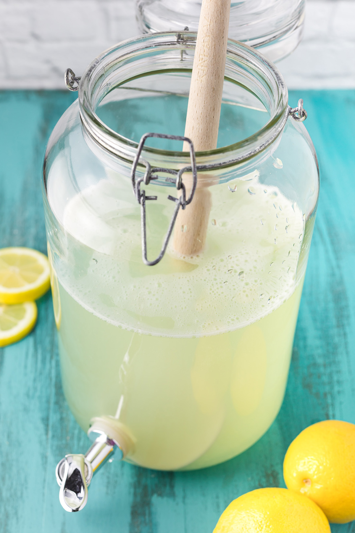 Stirring homemade hard lemonade in a glass dispenser with a wooden spoon.