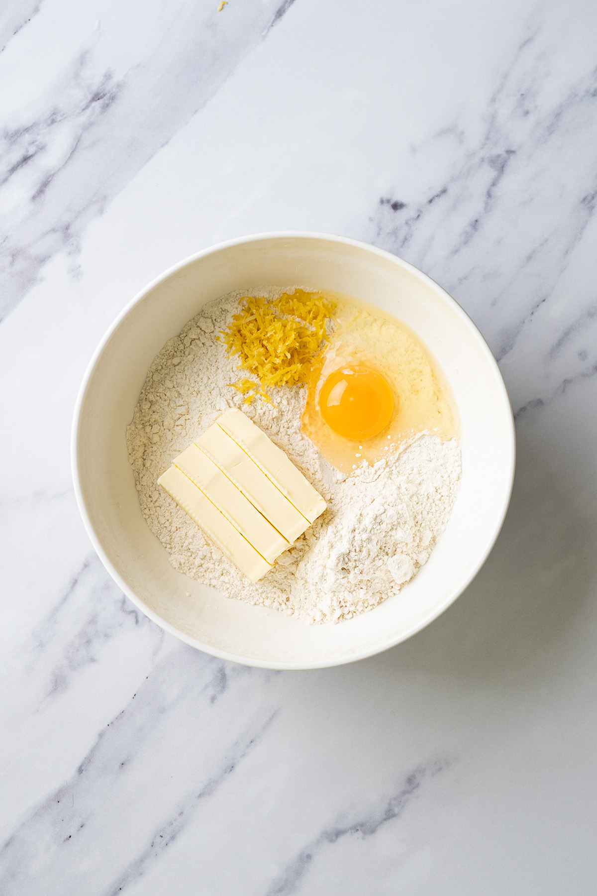 Dry ingredients, butter, egg, and fresh lemon zest in a white mixing bowl, ready to be mixed into lemon thumbprint cookie dough.