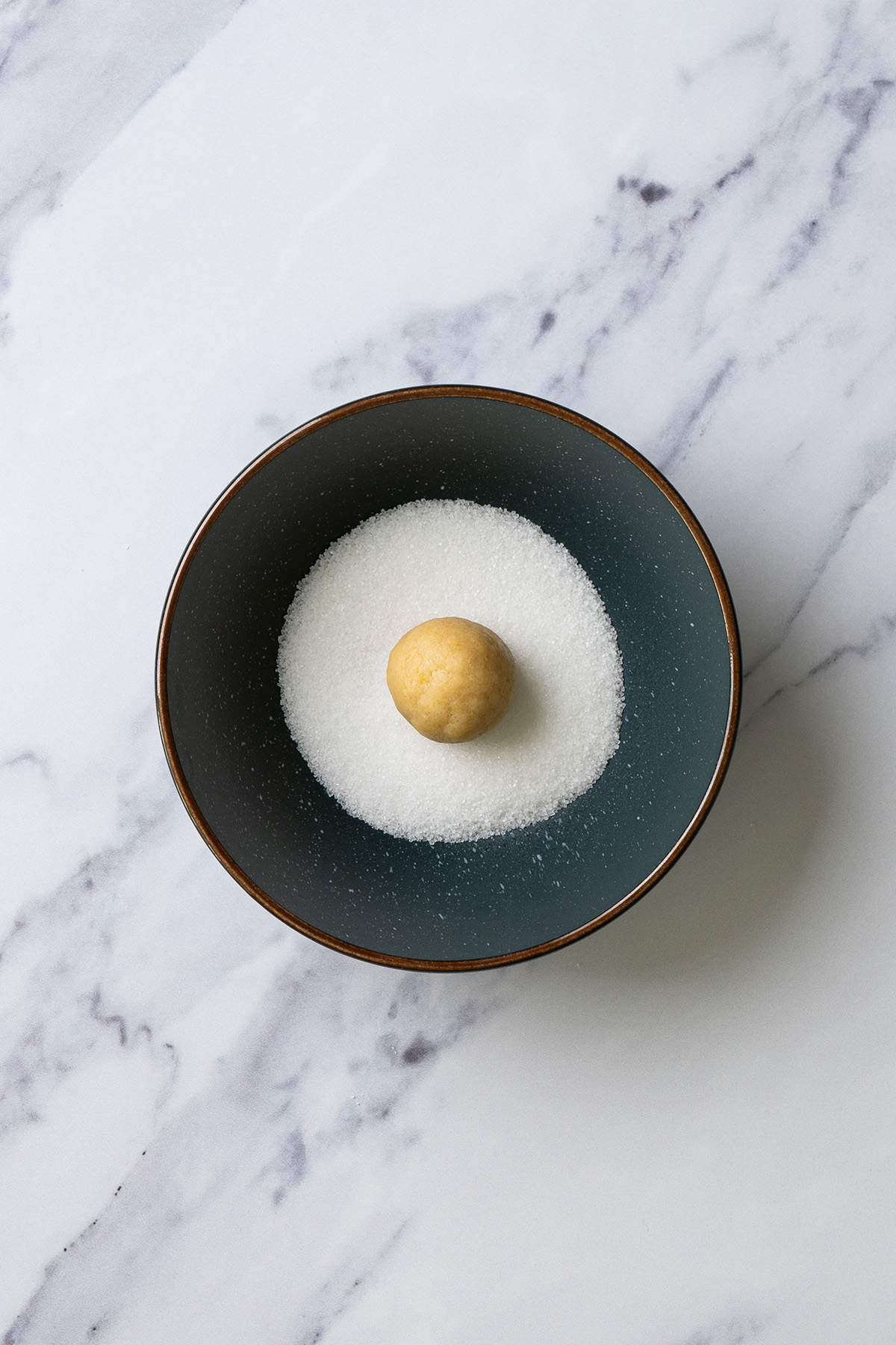 Cookie dough ball being rolled in granulated sugar in a small bowl before baking.