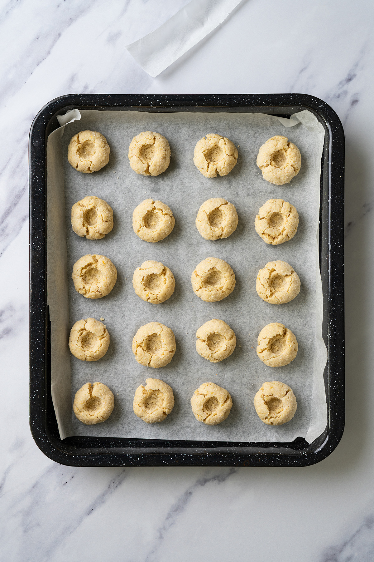 Freshly baked thumbprint cookies on a baking sheet with empty centers pressed into each cookie before filling.