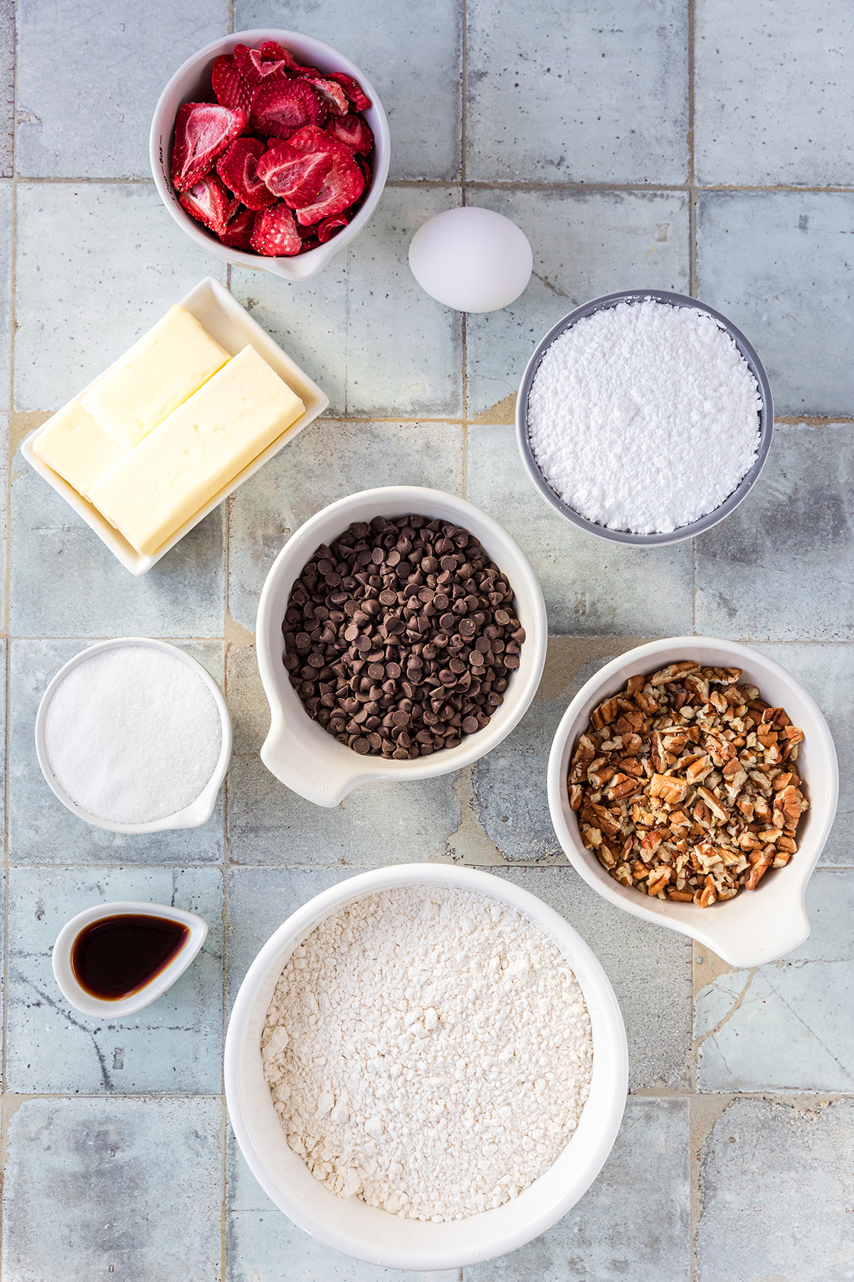 Flat lay of ingredients for strawberry snowball cookies including butter, flour, egg, sugar, powdered sugar, mini chocolate chips, chopped pecans, vanilla, and freeze-dried strawberries.